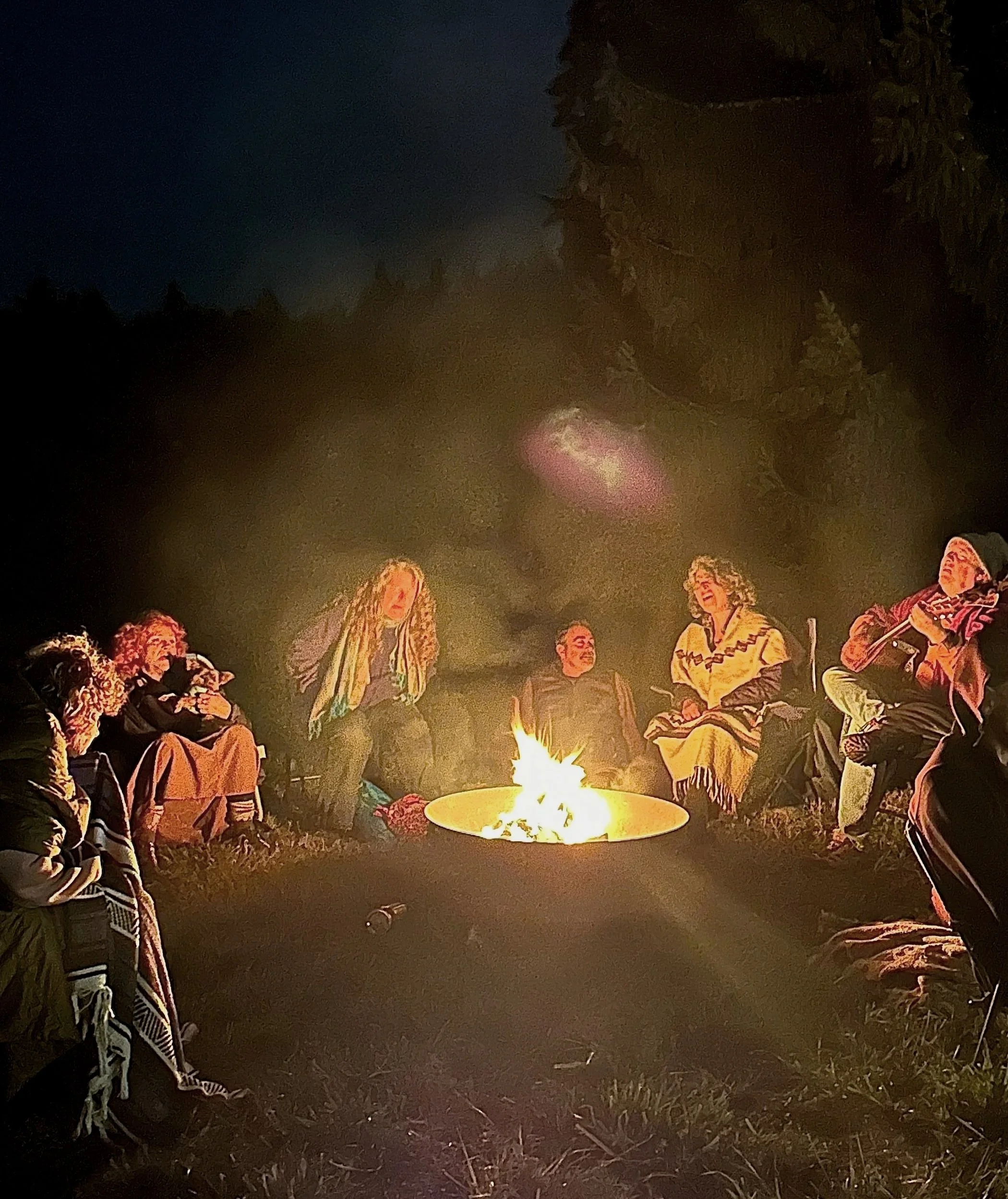 Group of people sitting around a campfire outdoors at night, surrounded by trees.
