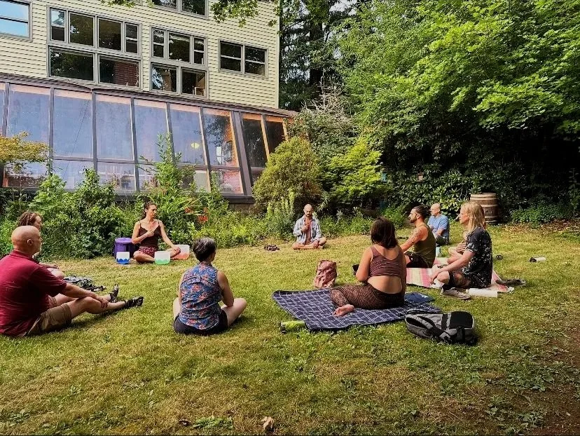 Group of people practicing yoga and meditation outdoors on a grassy area, surrounded by trees and bushes, near a building with large windows.