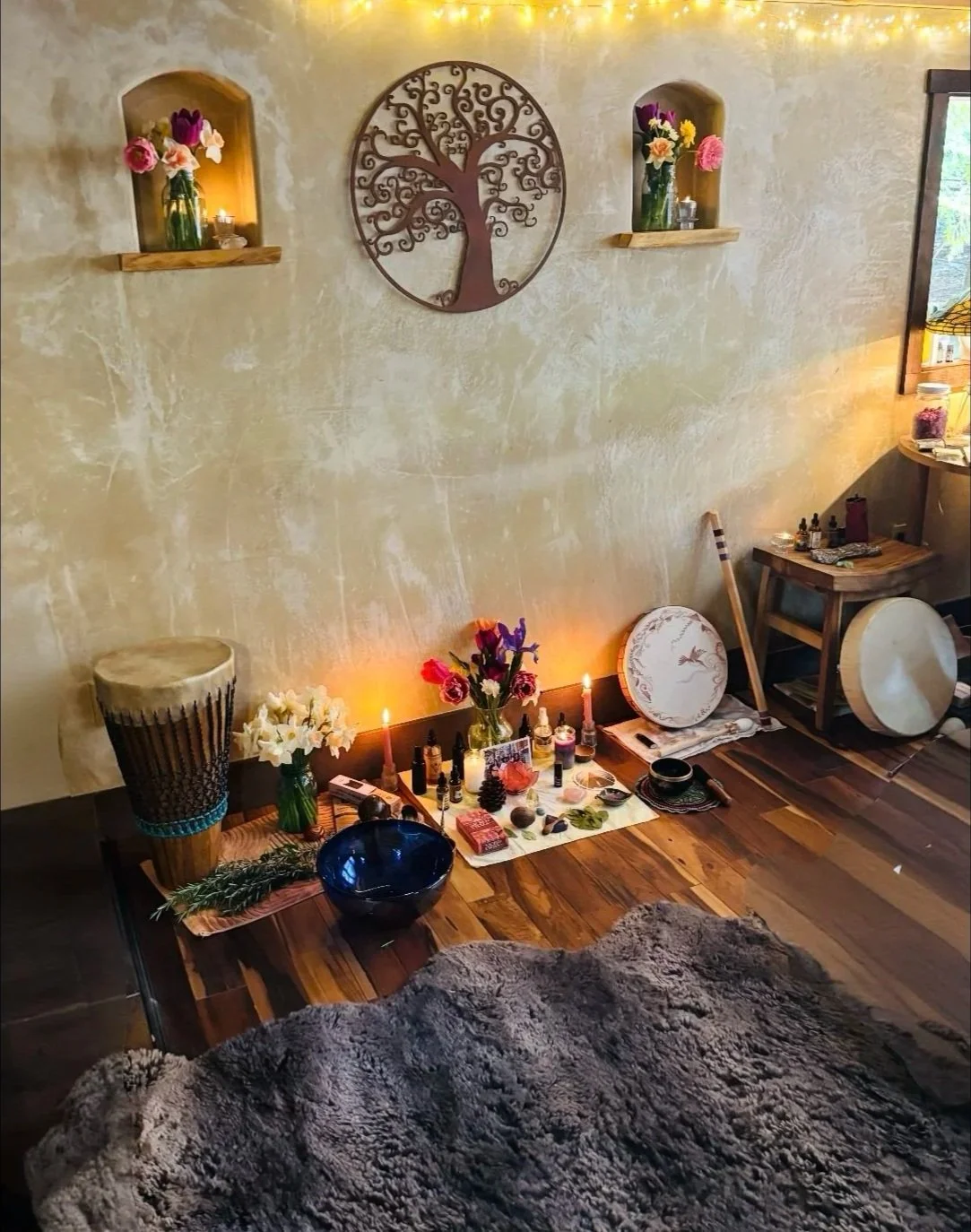 A cozy corner with spiritual and decorative items on a wooden floor, including flowers, candles, and ritual objects, against a textured beige wall with wall-mounted shelves, a circular wall art with a tree design, and bed and window on the right.