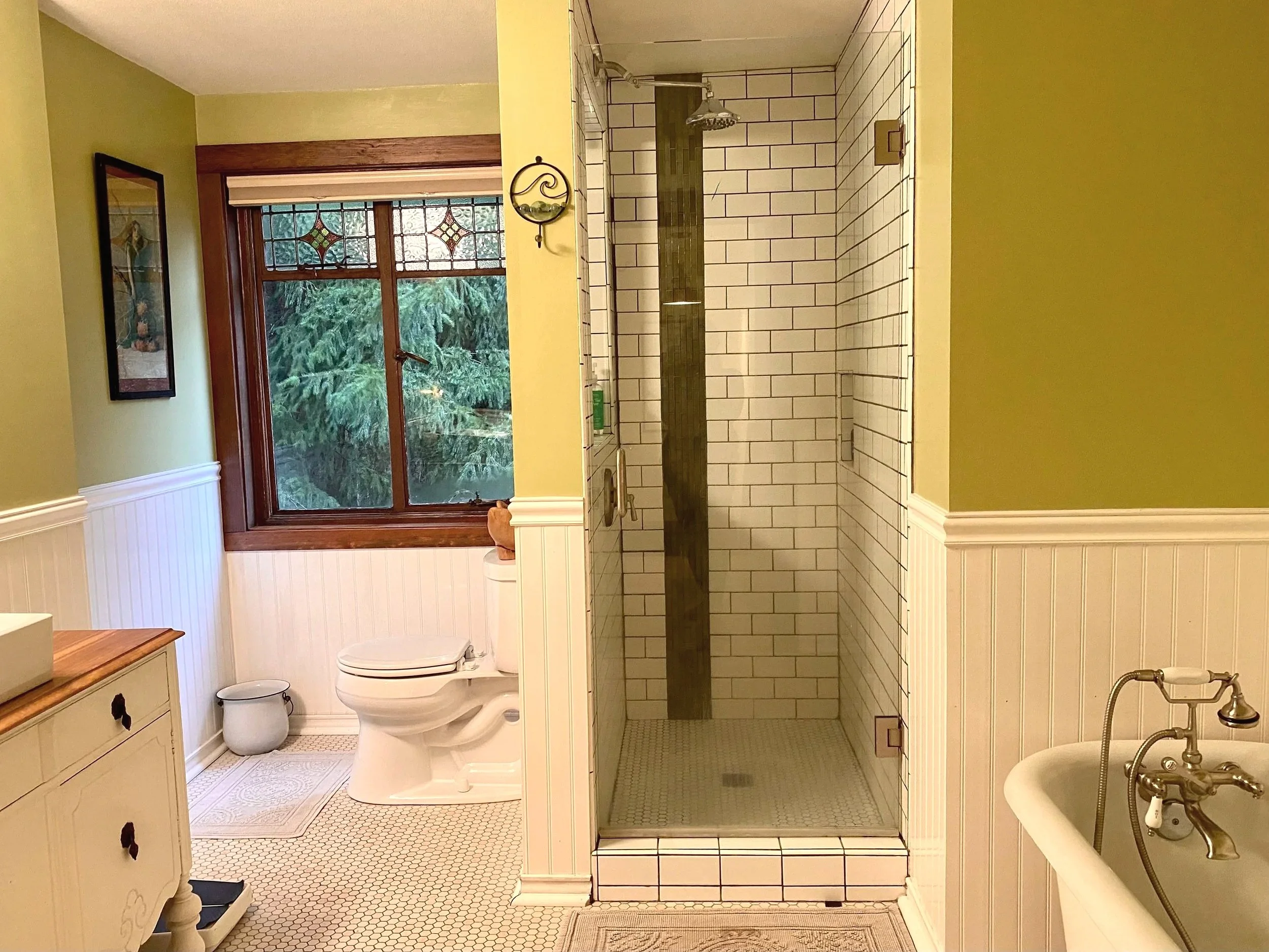 Bathroom with a large window, separate shower stall with white subway tiles and a dark accent tile, a white toilet, a wall-mounted mirror, a pedestal bathtub with a vintage-style faucet, and green walls with white wainscoting.