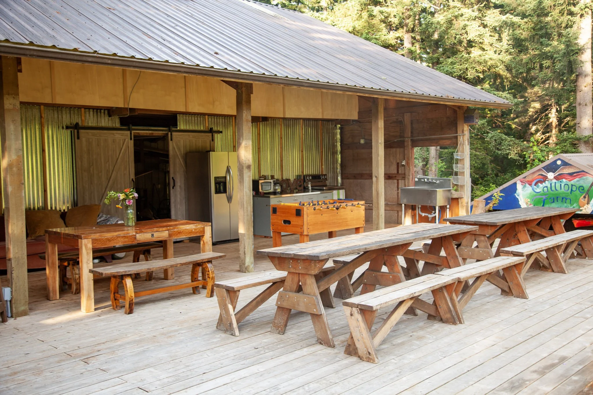 Outdoor patio with wooden picnic tables, a wooden table with a bench, an open cabin with a silver refrigerator, a foosball table, a sink, and a small corner with hanging baskets, surrounded by trees and greenery.