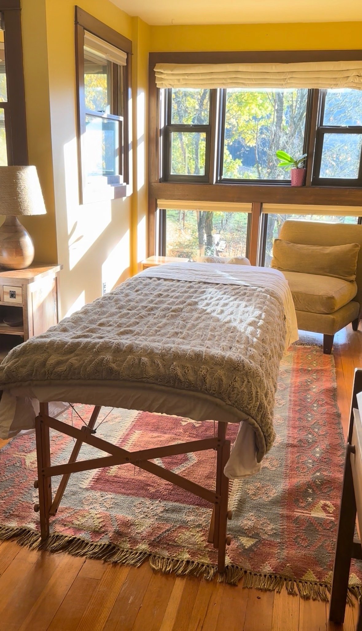 A cozy room with large windows showing trees outside, featuring a massage table with a plush blanket, a wooden nightstand with a lamp, a mustard yellow armchair, and a colorful rug on a wooden floor.