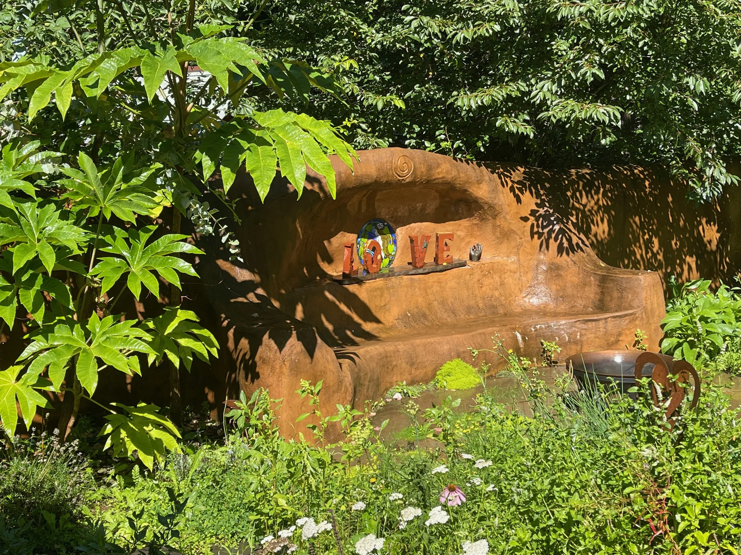 Garden scene with a large carved wood bench displaying decorative letters spelling 'LOVE', surrounded by green plants and flowers.