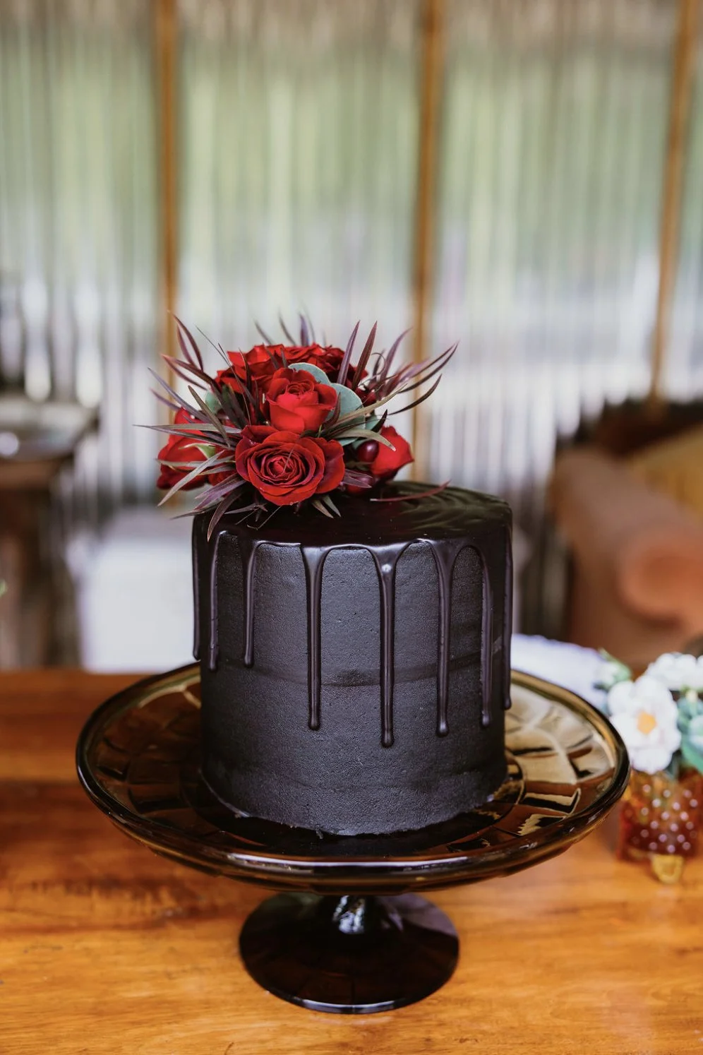 A black cake with chocolate drip decoration and a bouquet of red roses and dark leaves on top, displayed on a black cake stand on a wooden table.