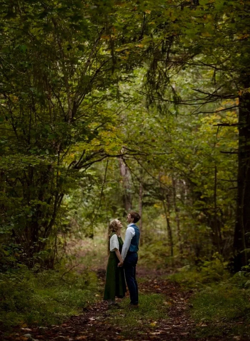 A man and woman dressed in vintage clothing stand close together in a forested path, holding hands and gazing at each other.