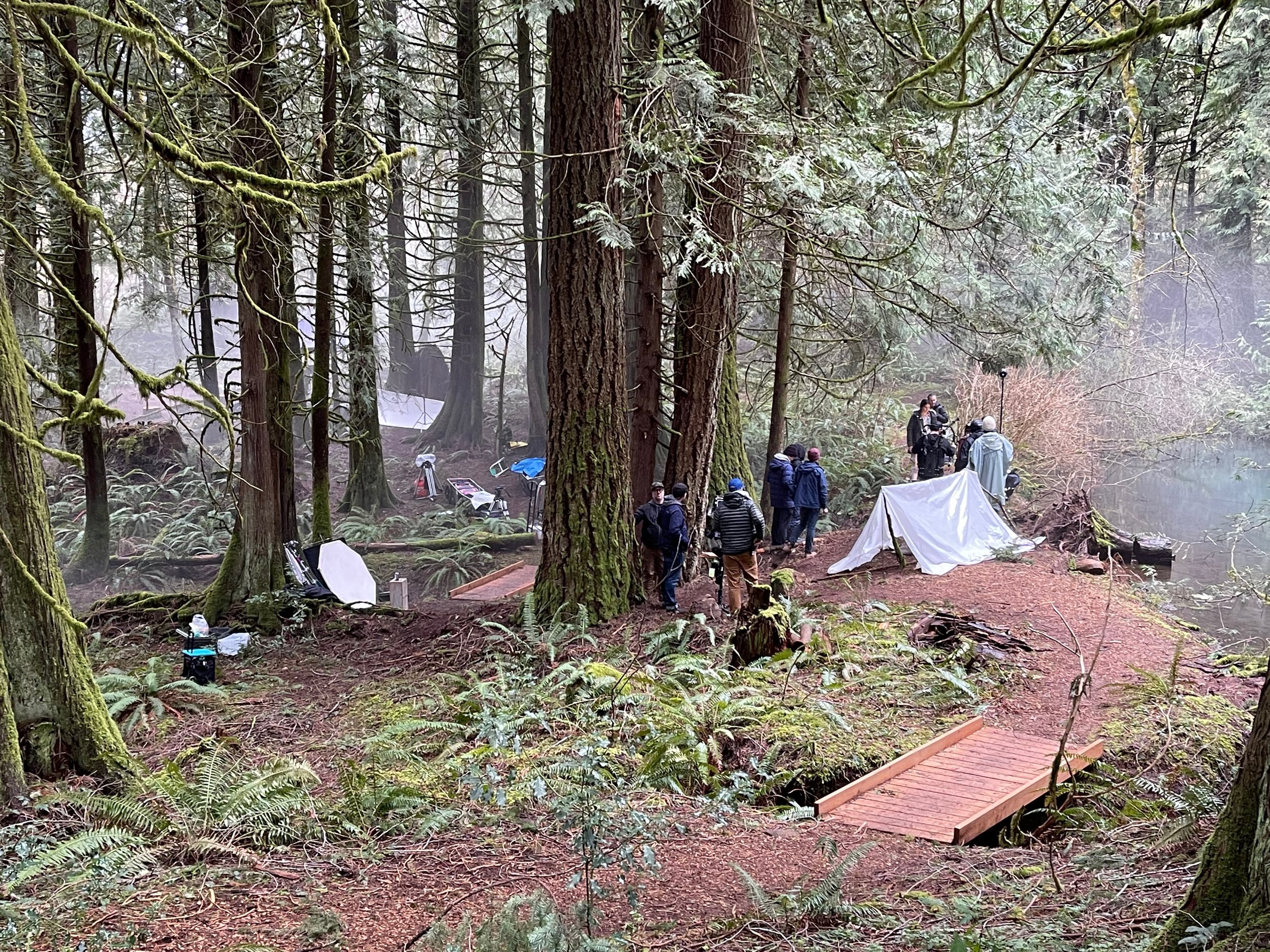 Film crew setting up equipment in a forest with tall trees and moss covering the trunks, with a small wooden bridge in the foreground, and a white tent and people in jackets gathered around in the background.