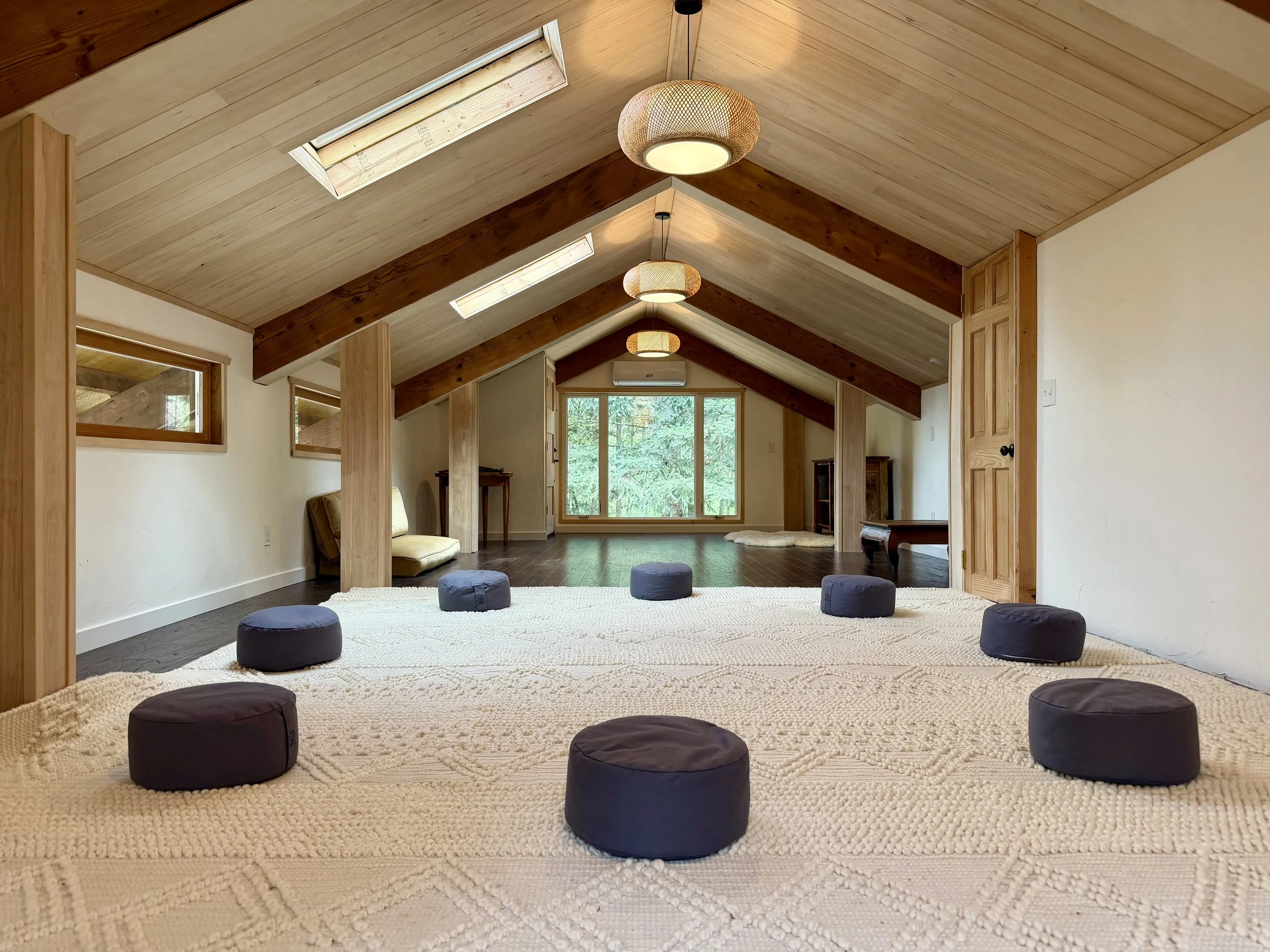 A cozy attic room with a sloped wooden ceiling, large windows, and a white carpeted floor with nine small black cushions arranged in a semi-circle.