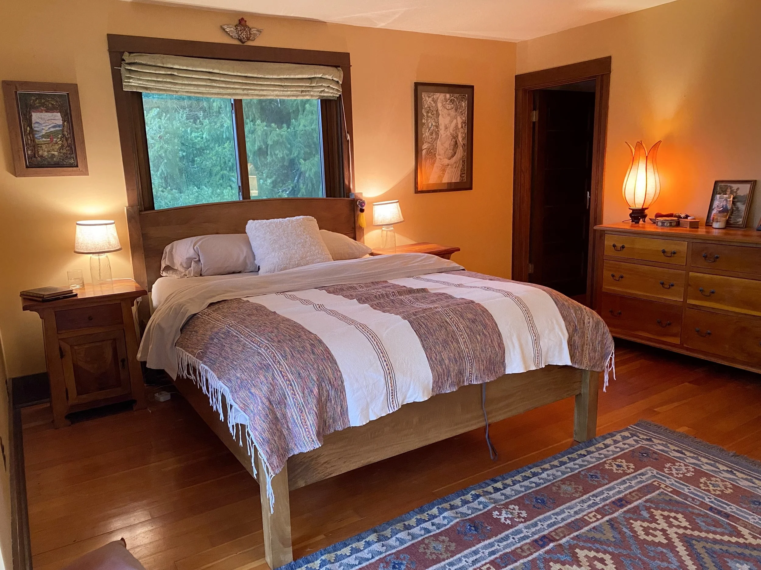 A cozy bedroom with warm lighting, a large bed with a wooden headboard and patterned quilt, two bedside lamps, a dresser with a lamp and framed photos, wooden flooring, and framed artwork on the walls.