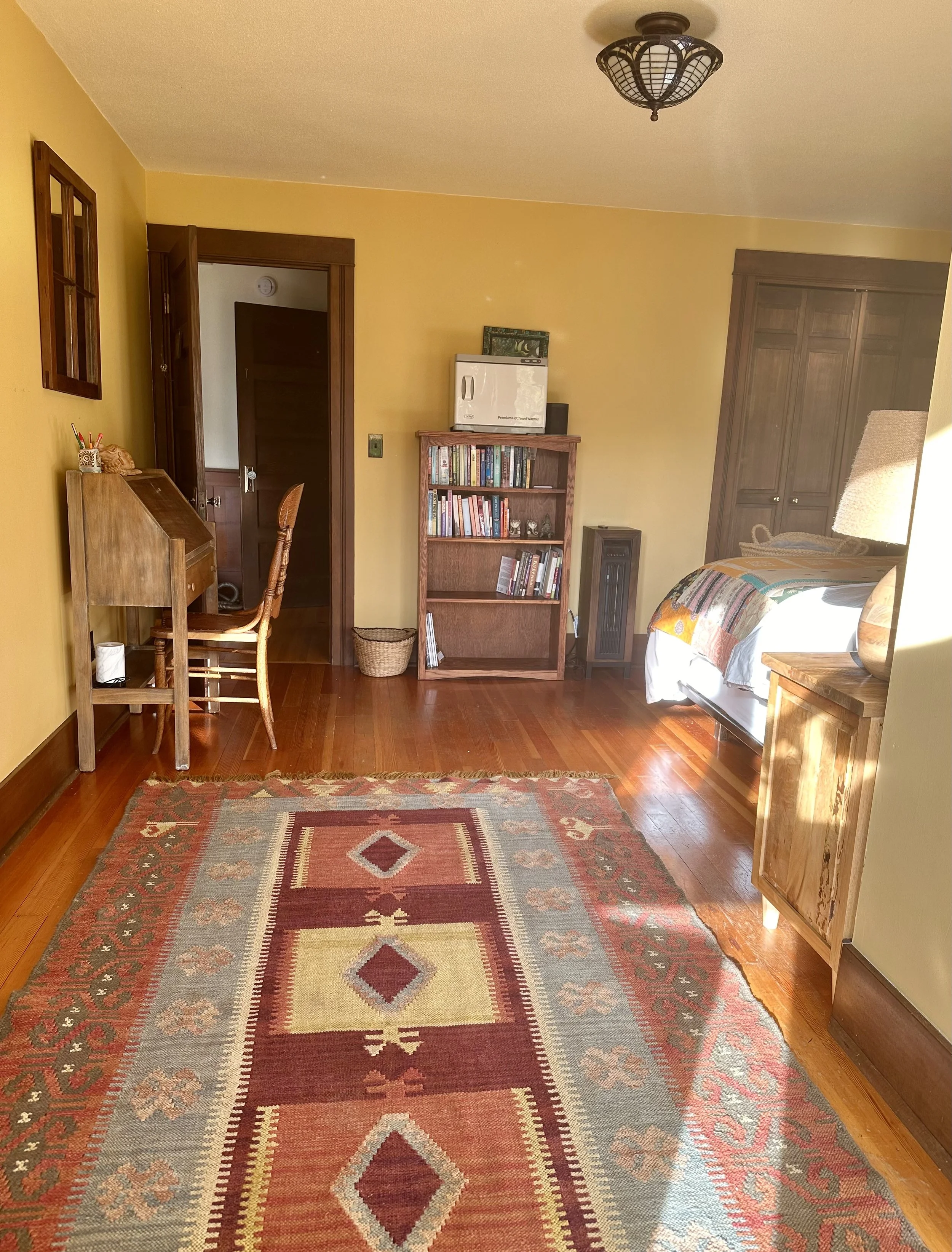 A cozy bedroom with yellow walls, hardwood floors, a colorful patterned rug, a wooden desk with a chair, a bookshelf filled with books, a bed with patchwork quilt, a wooden cabinet, and a ceiling light fixture.