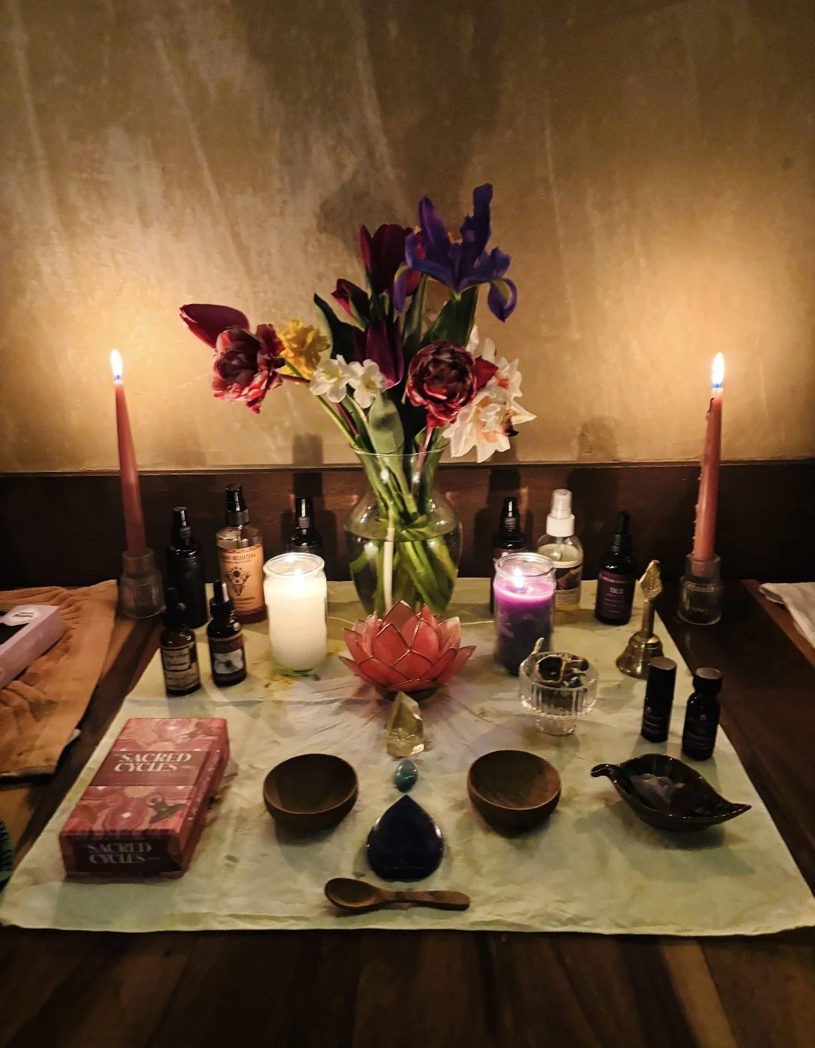 A spiritual altar with a vase of mixed flowers, lit candles, essential oils, and spiritual tools on a white cloth, set against a beige wall.