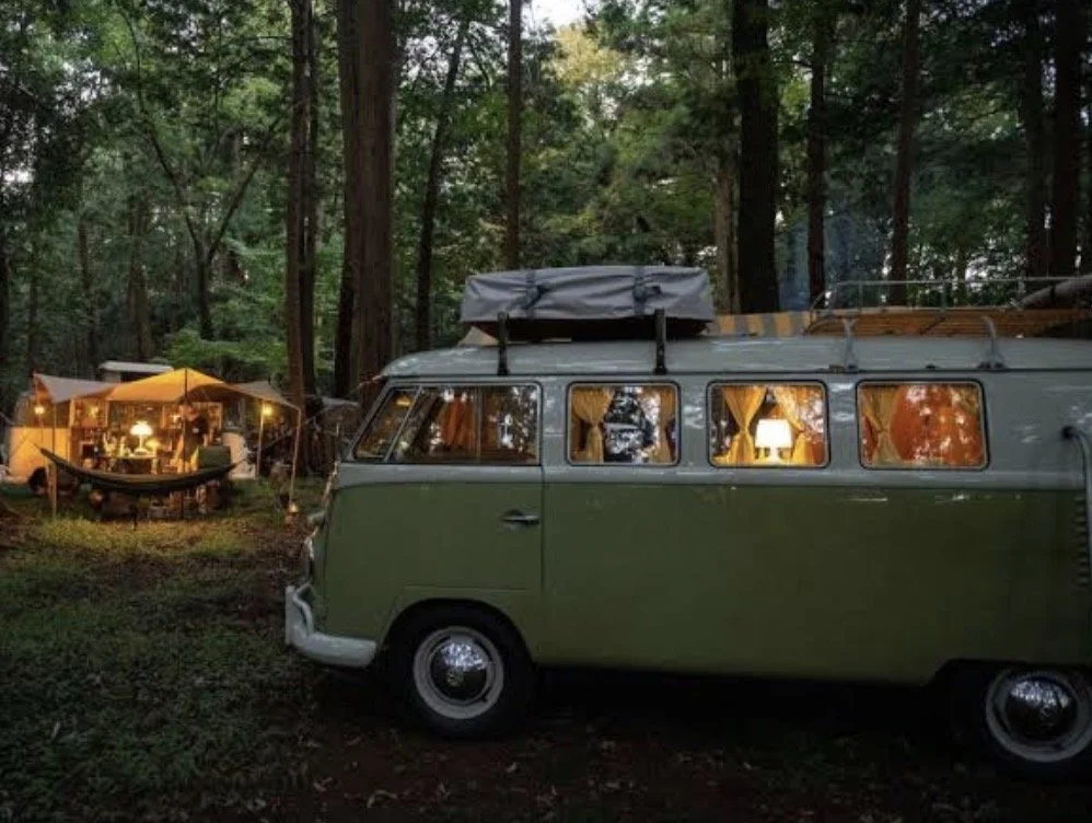 A vintage green van parked in a wooded campsite illuminated by warm interior lights and outdoor lamps, with a tent and hammock in the background.