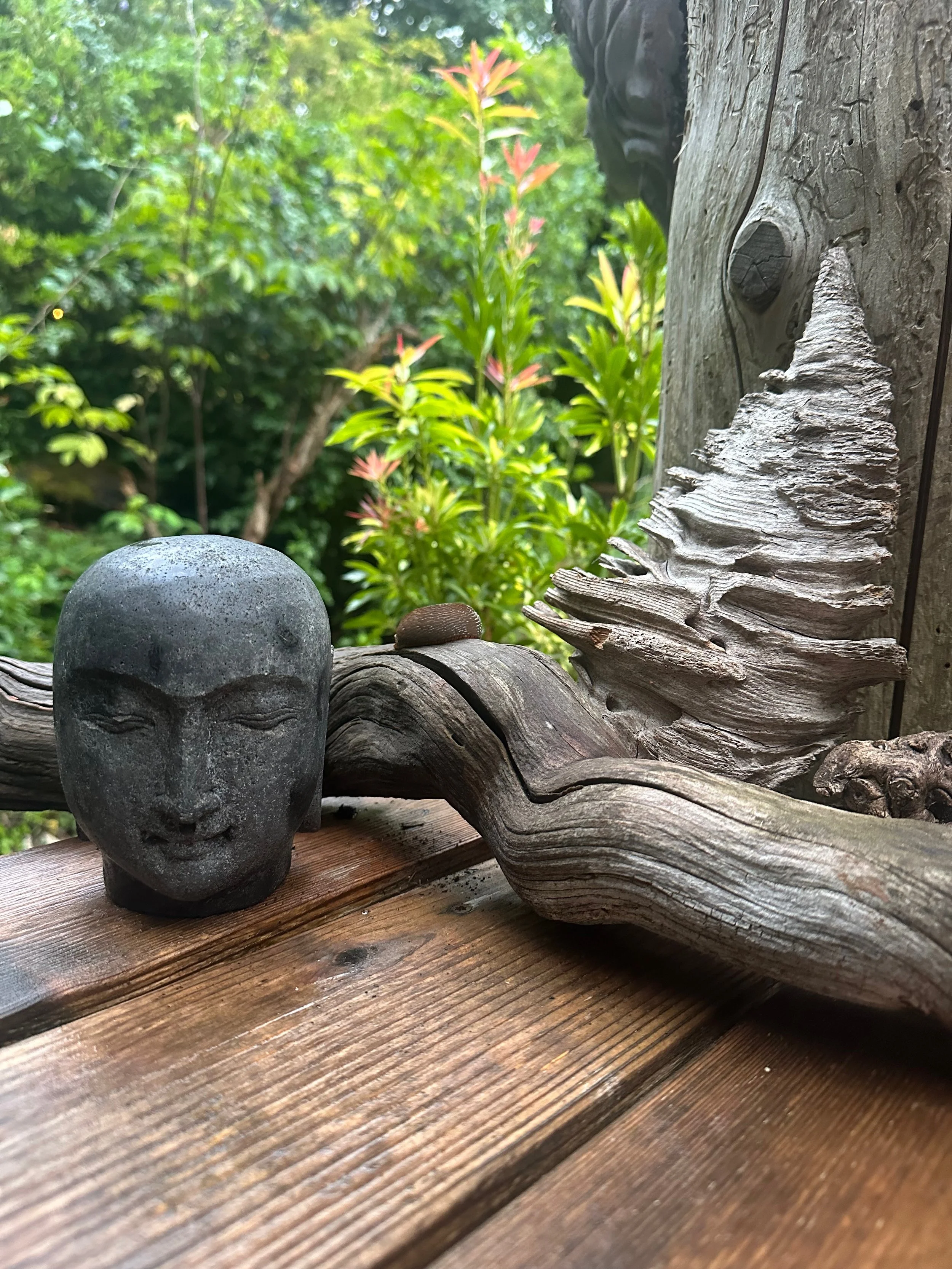 A sculpture of a face on a wooden surface with a twisted piece of driftwood and a conch shell beside it, outdoors with greenery in the background.