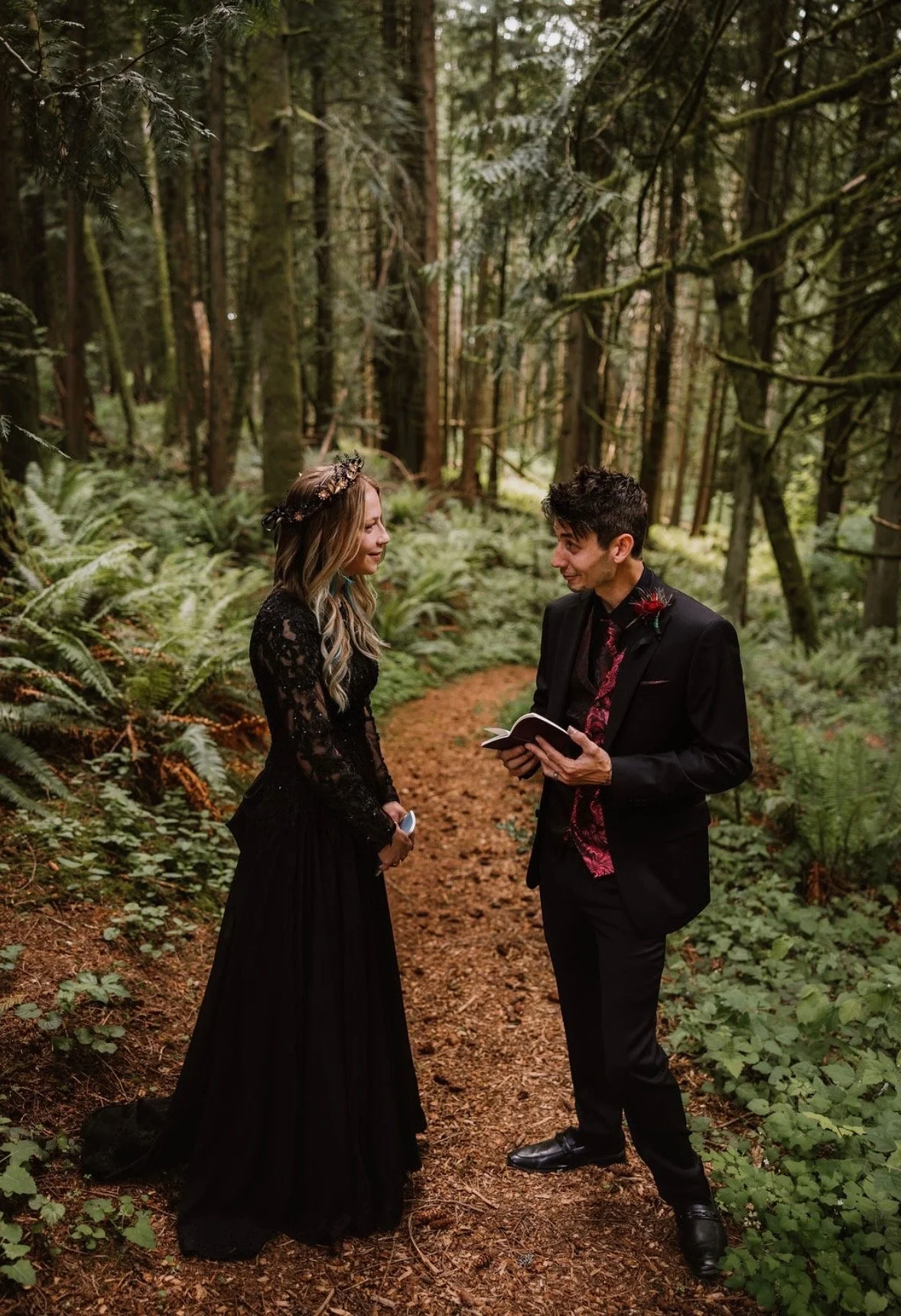 A couple dressed in formal black attire standing on a forest trail, with the man holding a small book or notebook and the woman looking at him, surrounded by lush green trees and plants.