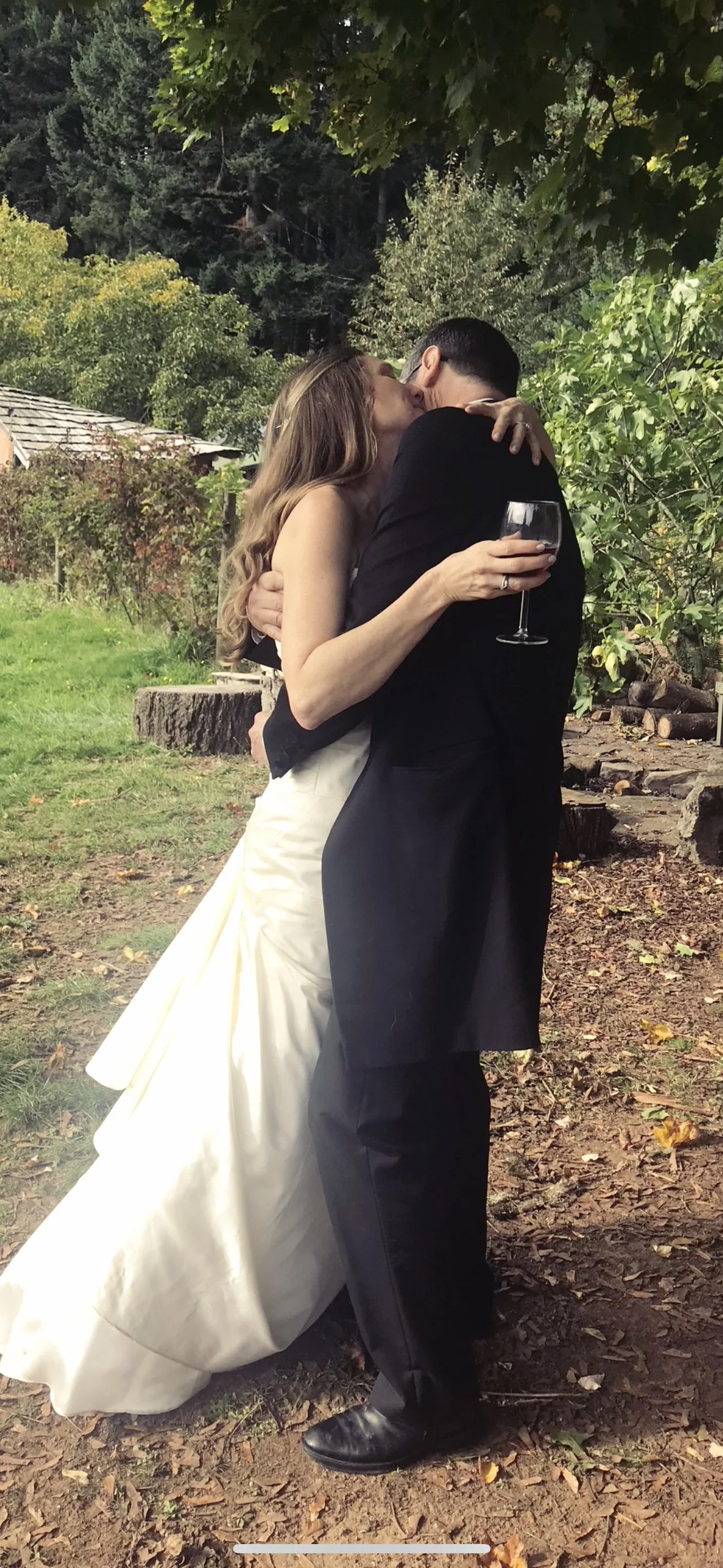 A bride and groom embracing and kissing outdoors, with the bride holding a glass of red wine, surrounded by green trees and fallen leaves.