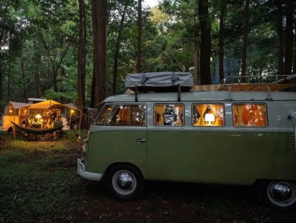 A green vintage camper van parked in a forest, with a lit outdoor camping setup with a hammock and chairs nearby.