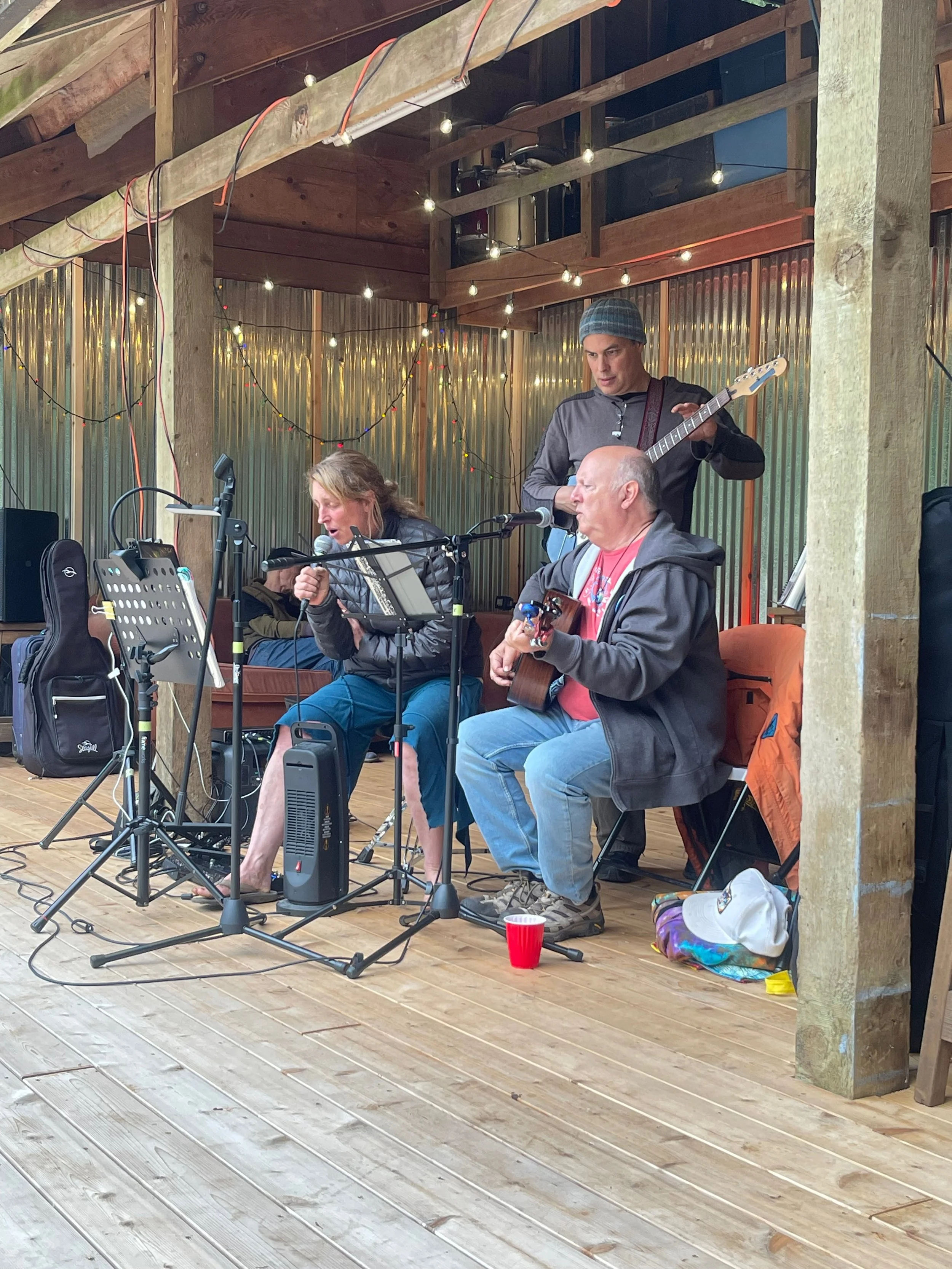 Three musicians performing on a wooden stage, with one playing guitar, another singing into a microphone, and a third playing a bass guitar, in a rustic setting decorated with string lights.