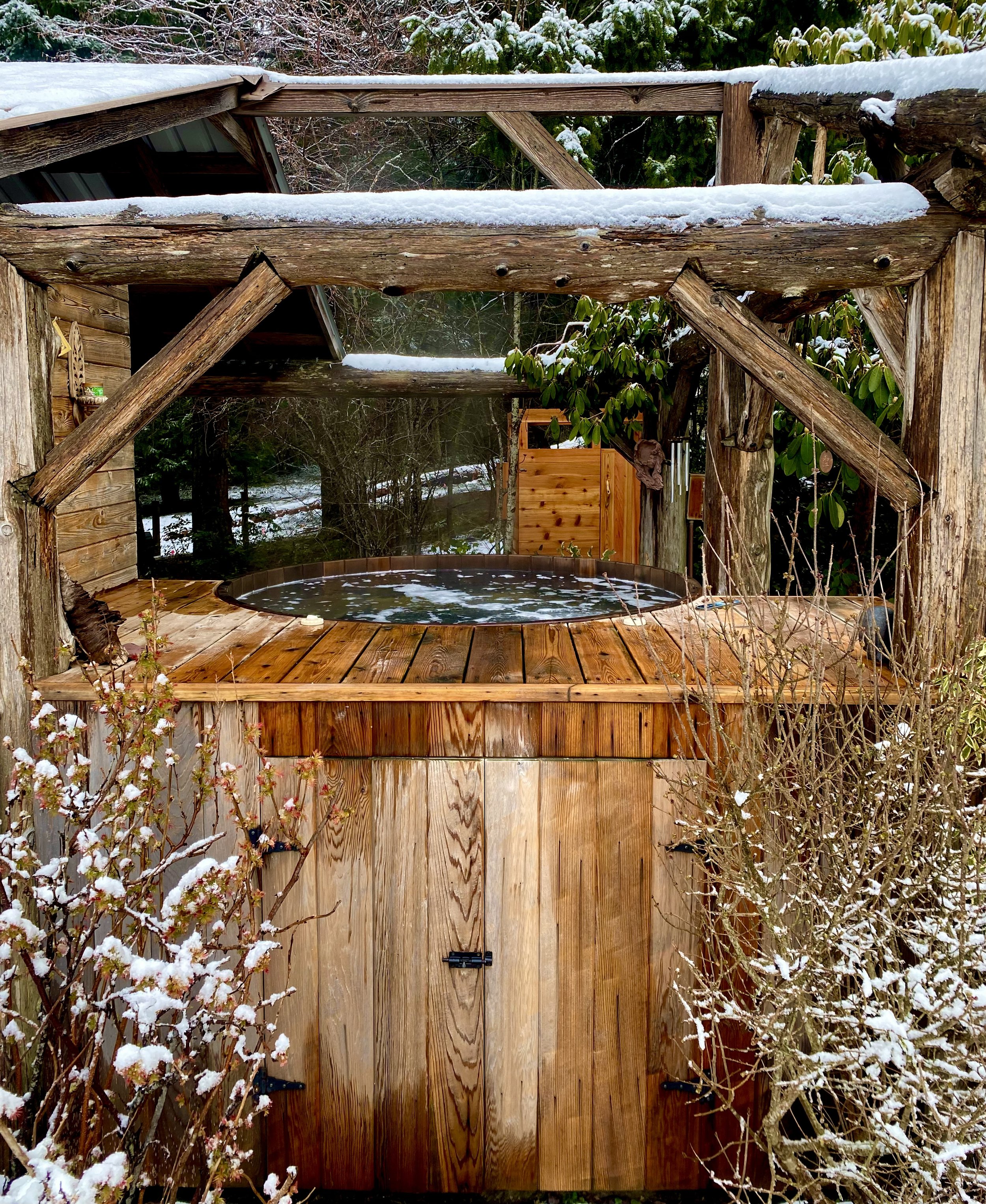 A wooden outdoor hot tub on a deck, surrounded by snow-covered bushes and trees, with a rustic wooden structure and a shed in the background.