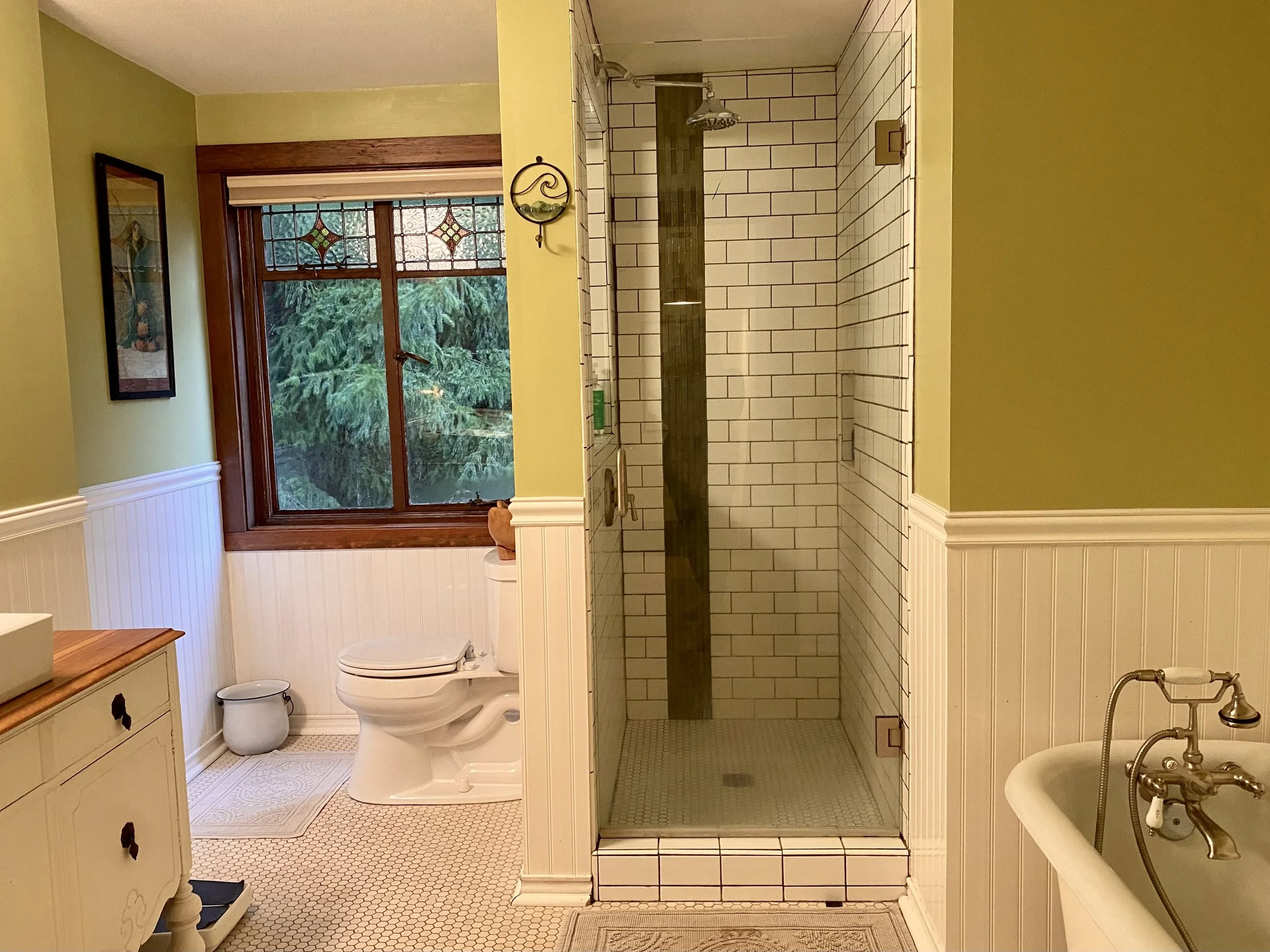 Bathroom with green walls, white wainscoting, wood-framed window with stained glass, toilet, shower with white subway tiles, and a vintage bathtub with brass faucet
