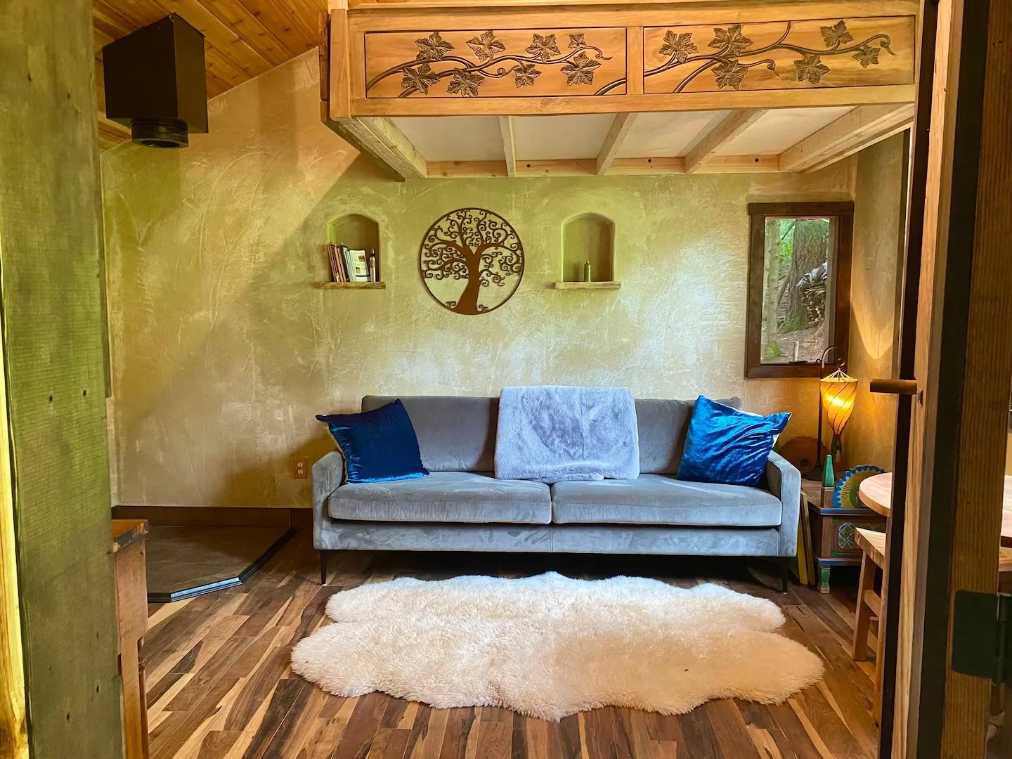 Living room with a gray sofa accented by two blue pillows, a white throw blanket, a white fluffy rug, a wooden side table, a small lamp, and a window showing greenery outside. The wall behind the sofa has two small shelves with books and decor, and a decorative metal tree wall art piece. There is a wooden loft or ceiling detail with carved vine and leaf pattern.