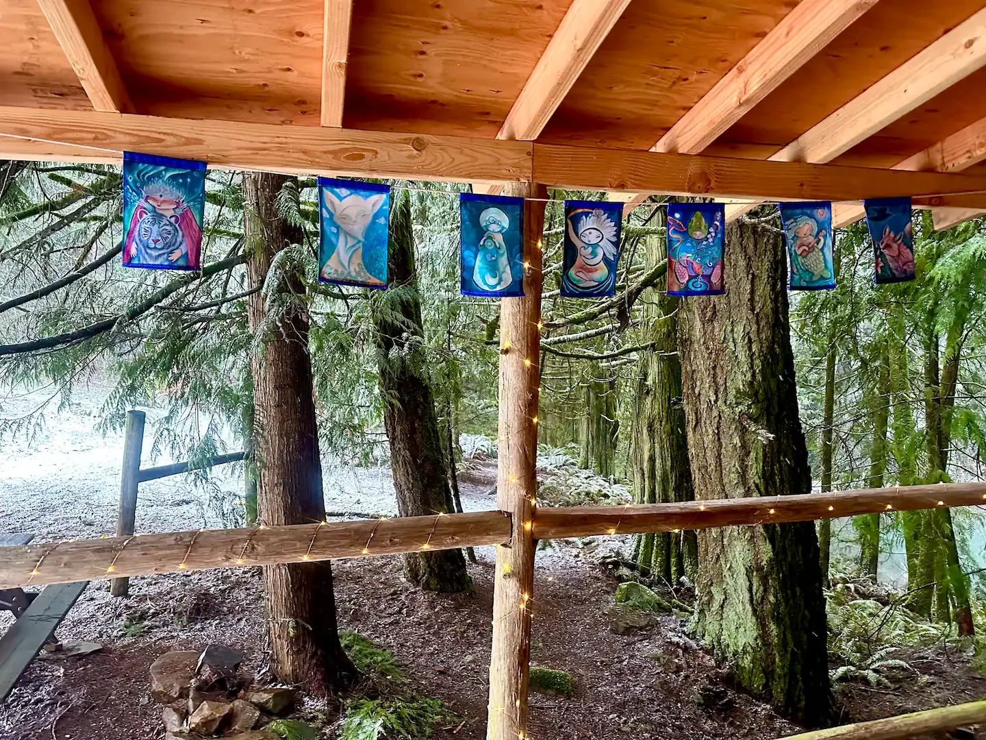 Colorful flags with mystical animal and character designs hang beneath a wooden structure in a forest setting, decorated with string lights.