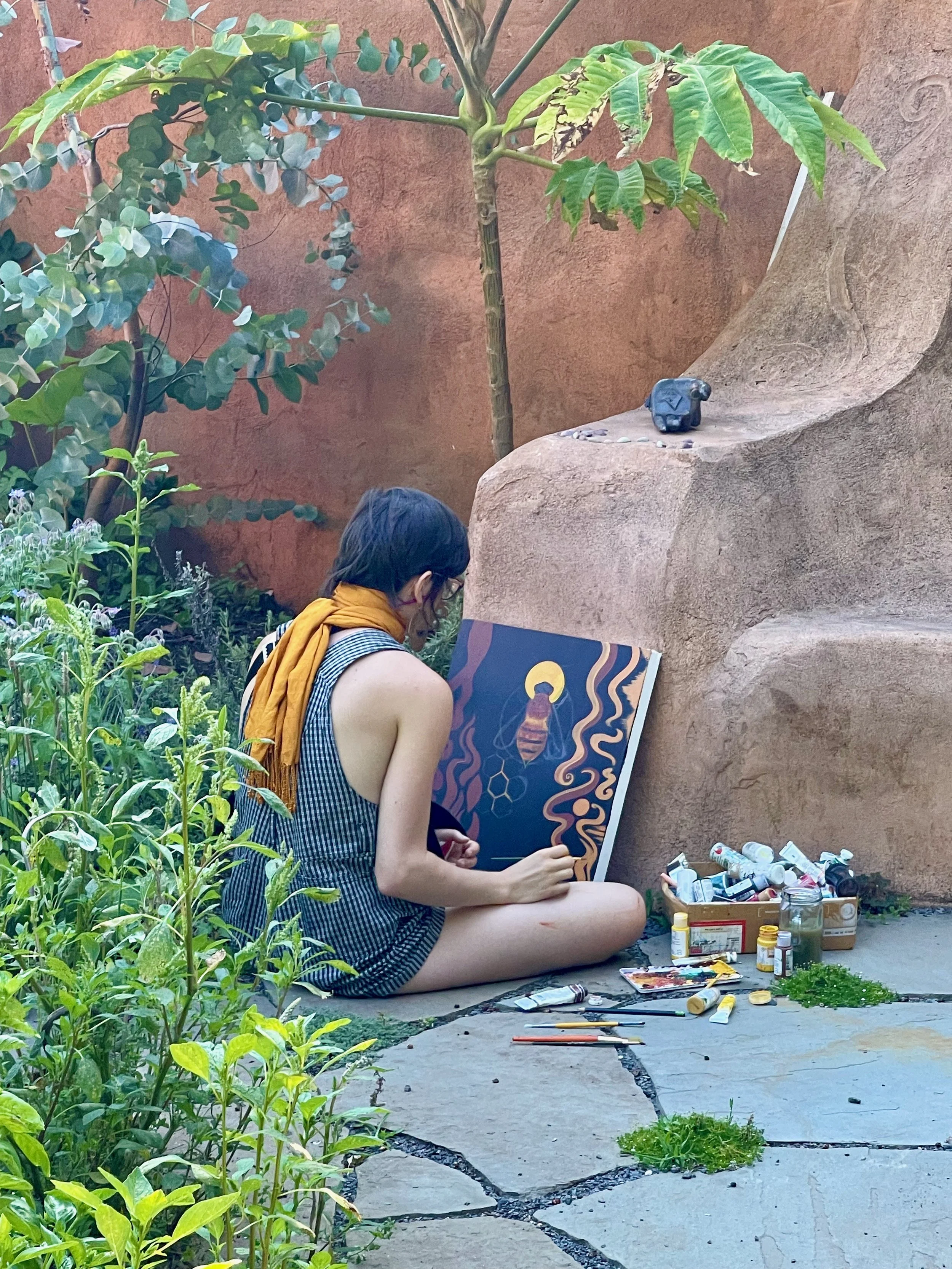 A person sitting on the ground outdoors, painting a colorful picture of a bee with abstract patterns on a canvas. The person has short dark hair, wears glasses, a sleeveless striped dress, and has a yellow scarf around their neck. They are surrounded