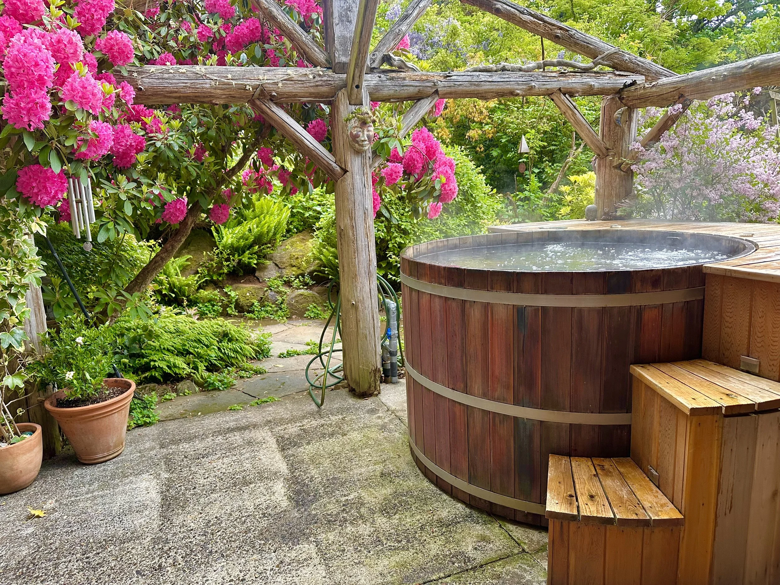 A wooden hot tub on a patio surrounded by pink and purple flowering bushes and green plants, with a wooden pergola overhead and a garden hose nearby.