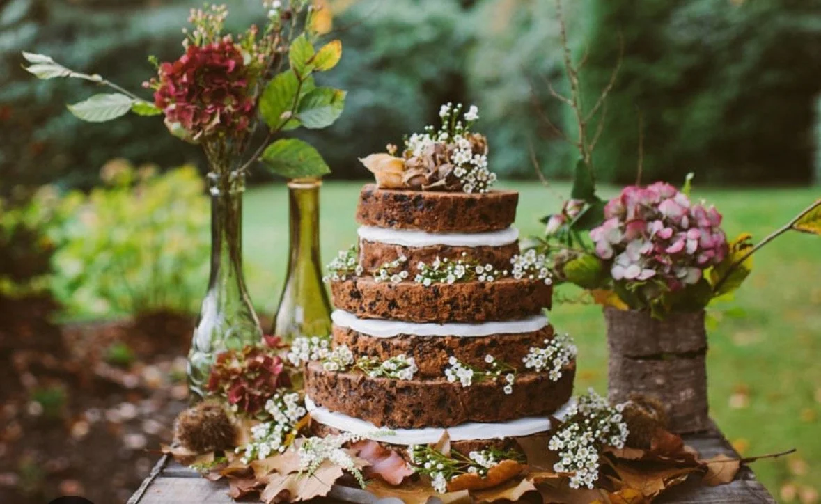 A rustic, four-layer chocolate cake with white cream between layers, decorated with small white flowers and dried leaves, surrounded by flowered vases and autumn leaves outdoors.