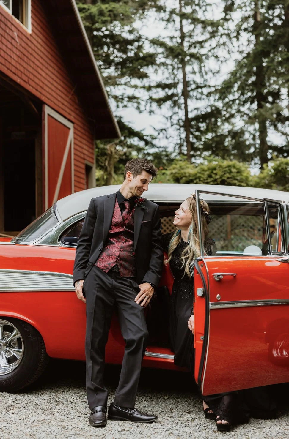 A man in a black suit and a woman in a black dress standing beside a vintage red and white car, smiling at each other outside a red barn surrounded by trees.