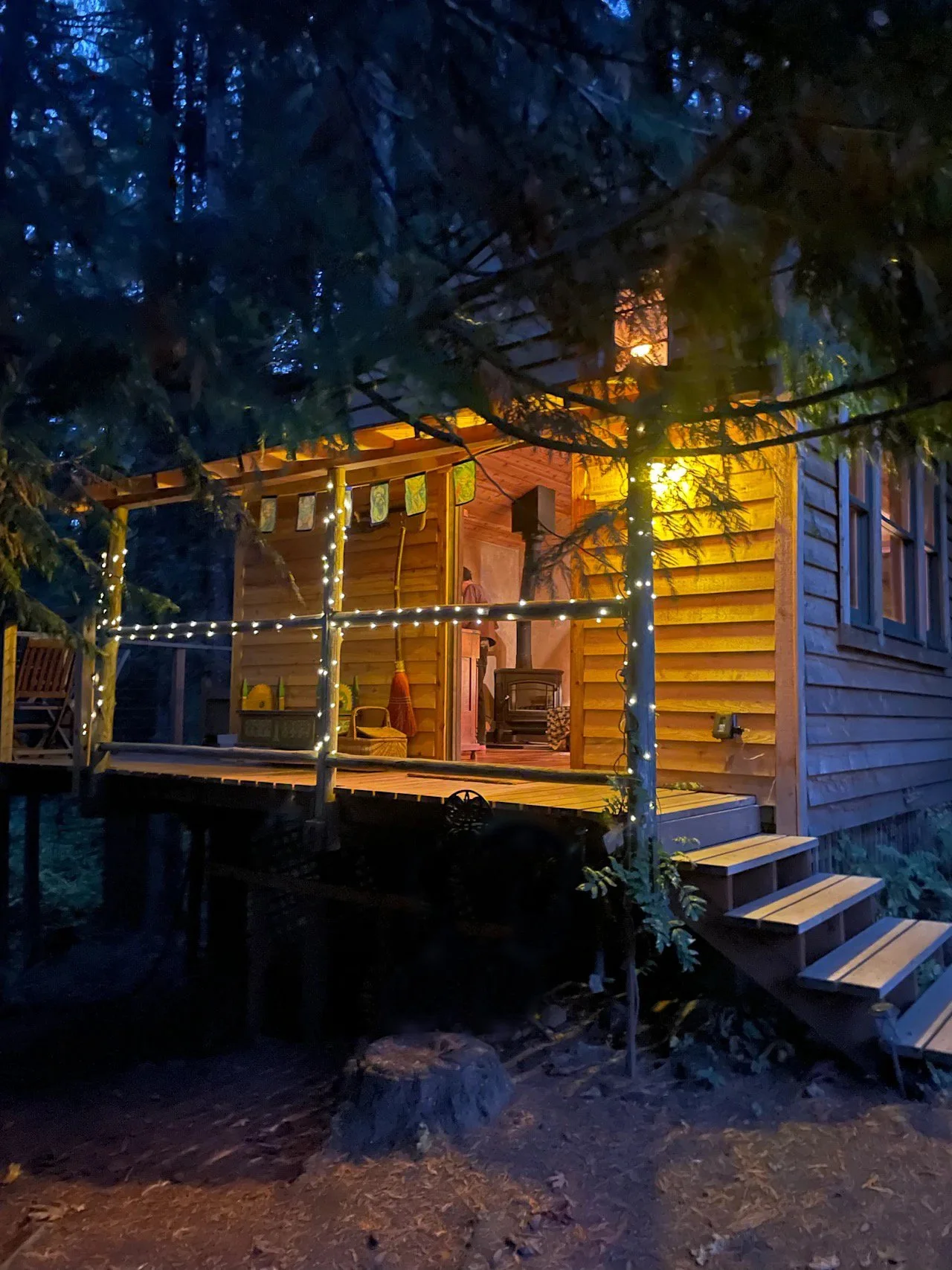 A wooden cabin illuminated with warm yellow lights is surrounded by trees. The cabin has a small porch with string lights, a pizza oven, and some chairs, all set in a forest at dusk or night.