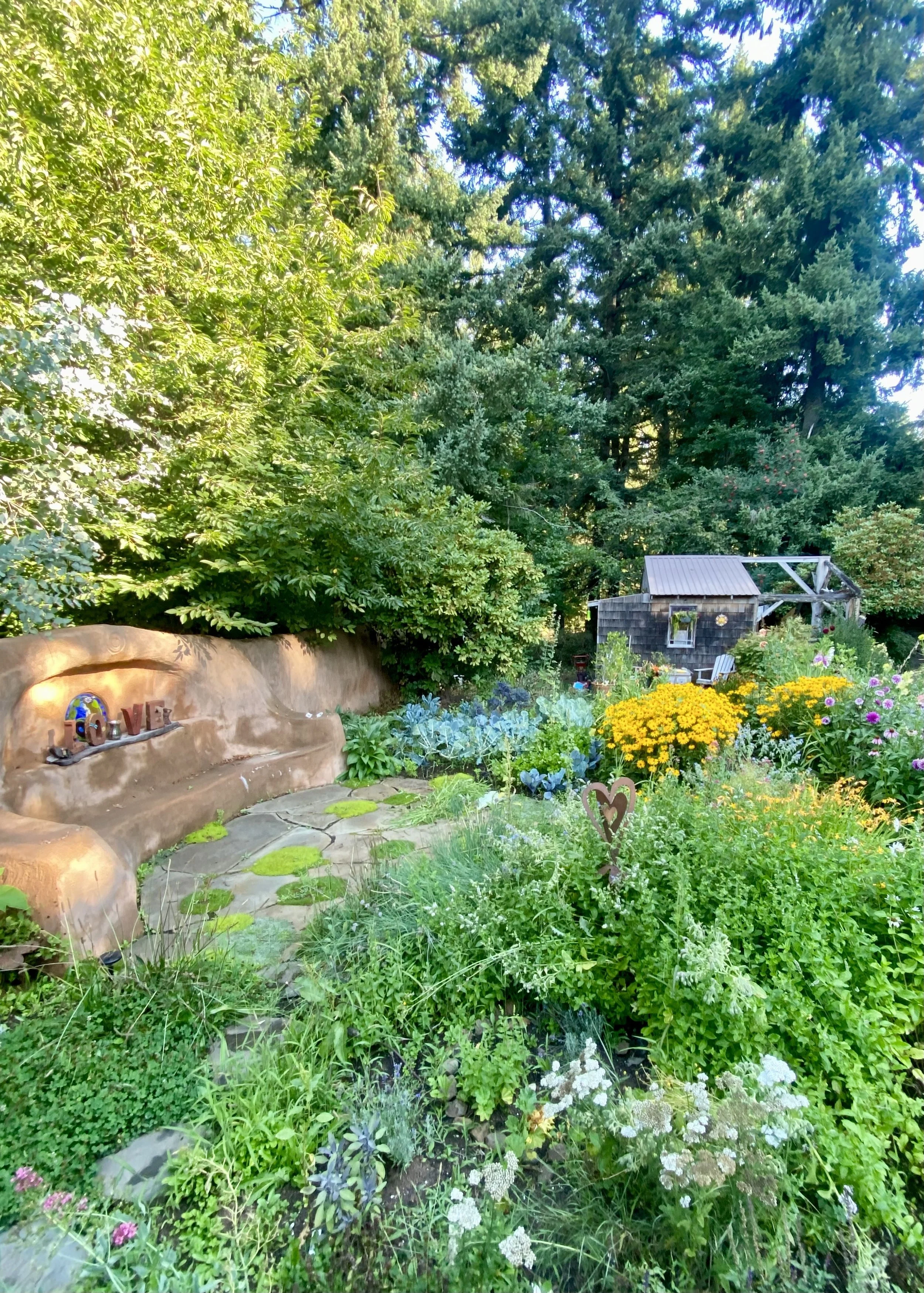 A lush garden with a variety of vibrant flowers and greenery, a rustic wooden shed, and a curved stone bench with decorative items, surrounded by tall trees.