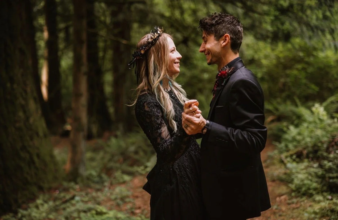 A couple in wedding attire dancing and smiling in a forest setting.