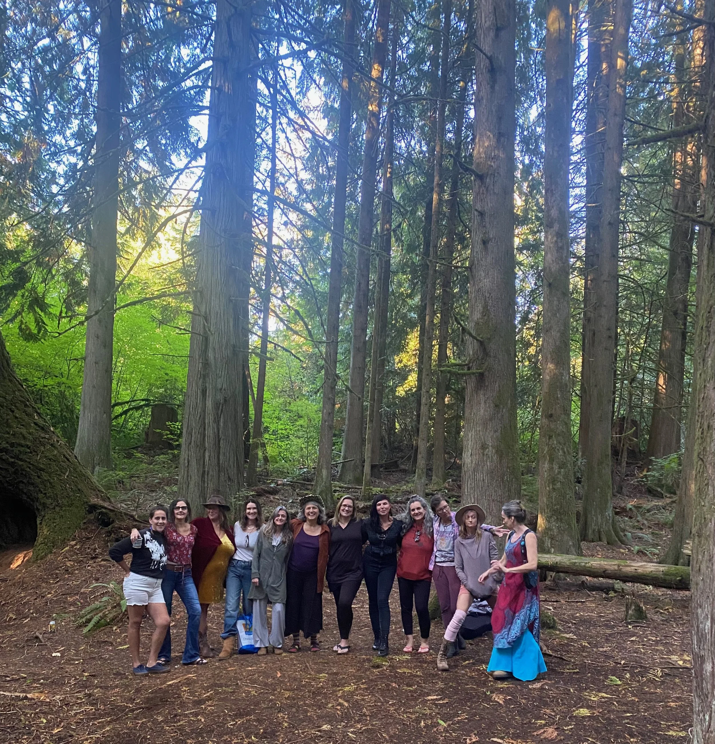 Group of women standing together in a forest, surrounded by tall trees and greenery.