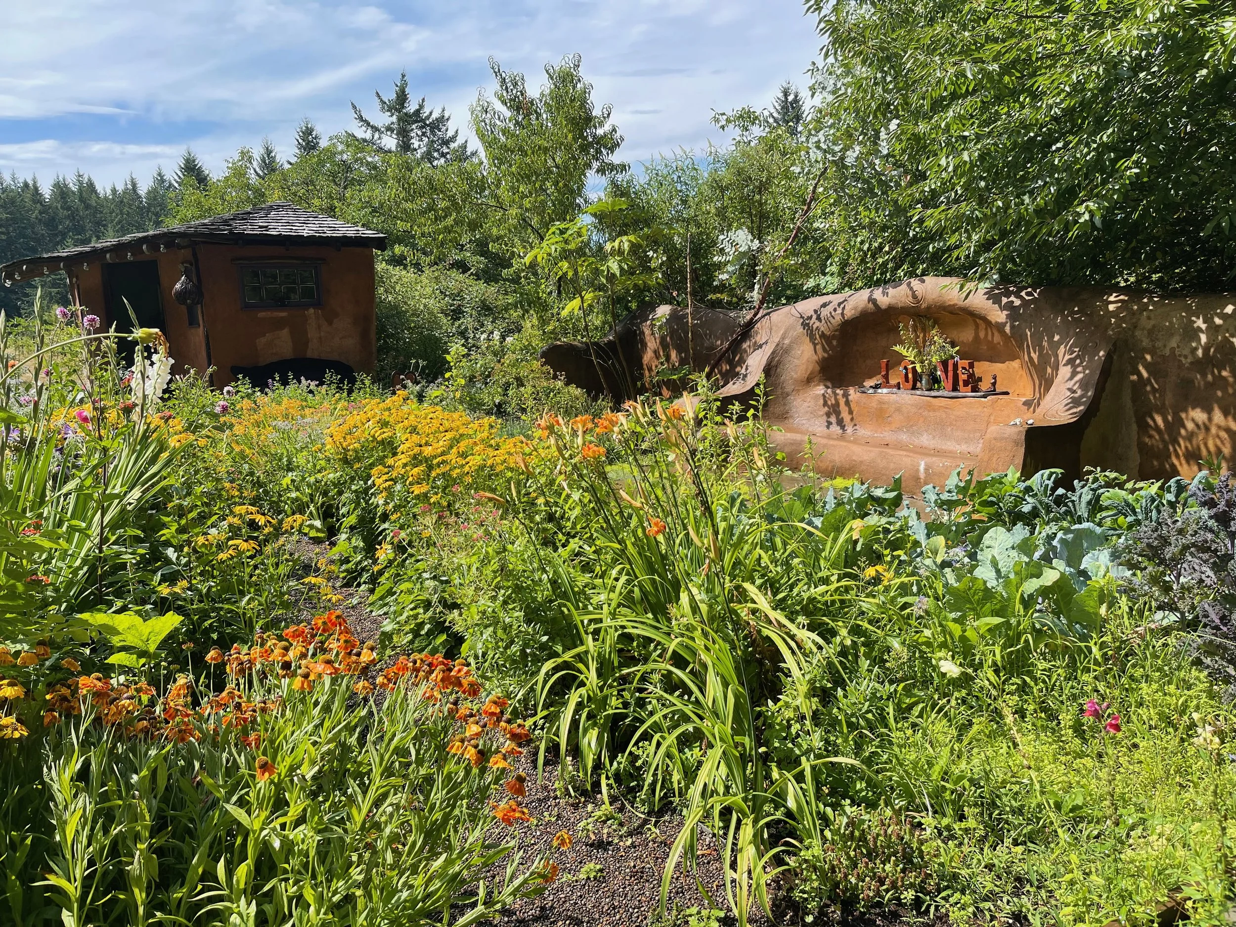 A lush garden with various colorful flowers and green plants, a small rustic wooden shed, and a curved wall with a shelf displaying decorative items spelling 'LOVE', under a blue sky with some clouds.