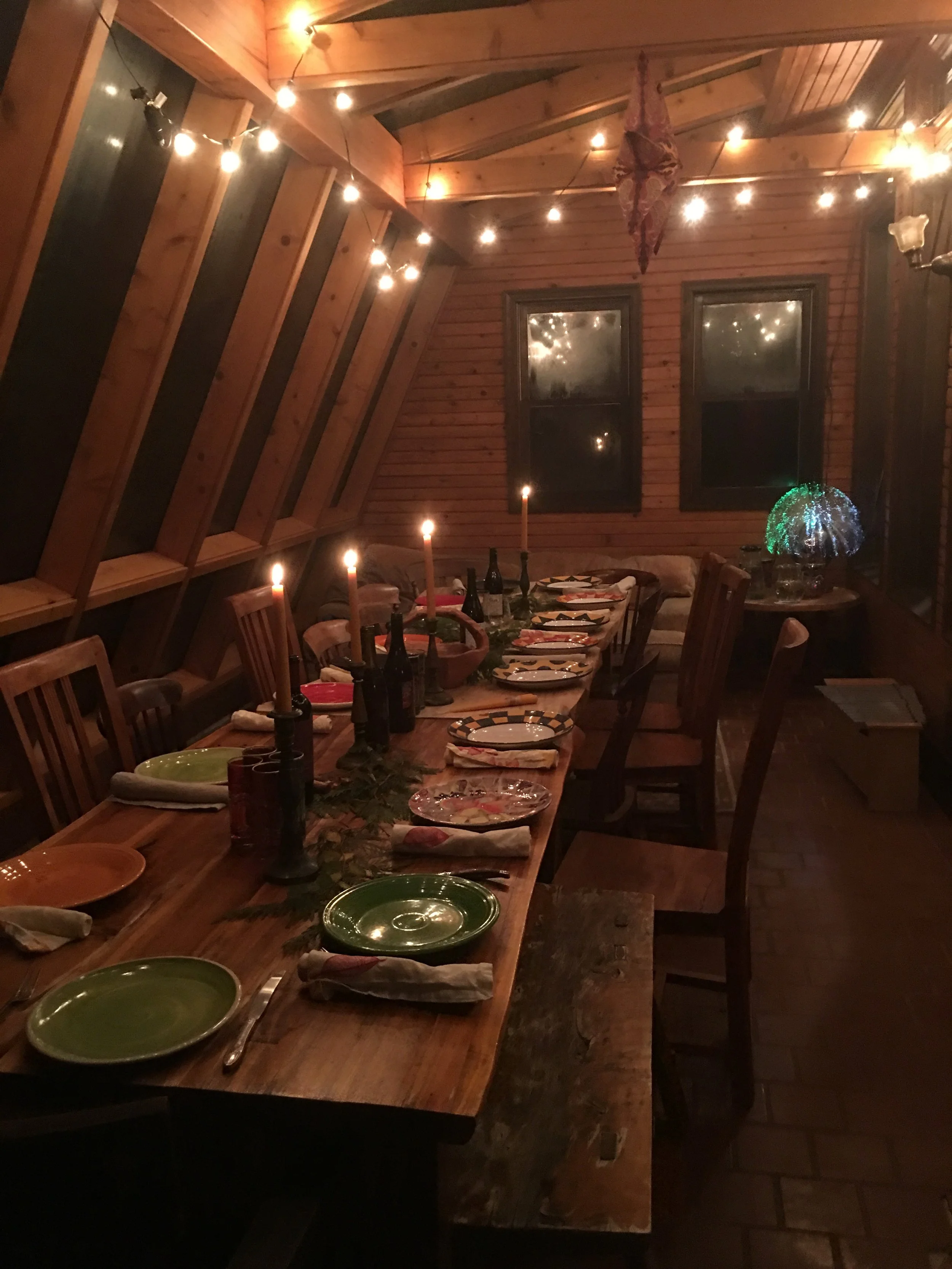 A warmly lit, rustic dining room with a long wooden table set for a holiday meal, featuring green and festive plates, napkins, and candles, with string lights and windows in the background.