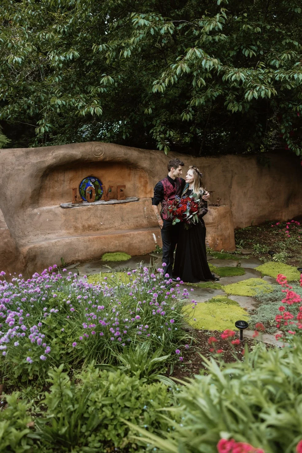 A couple standing in a garden, embracing each other, with a large rock formation in the background that spells out "LOVE" with a globe in the letter "O". The woman is holding a bouquet of red flowers, and the scene is surrounded by blooming pink and 