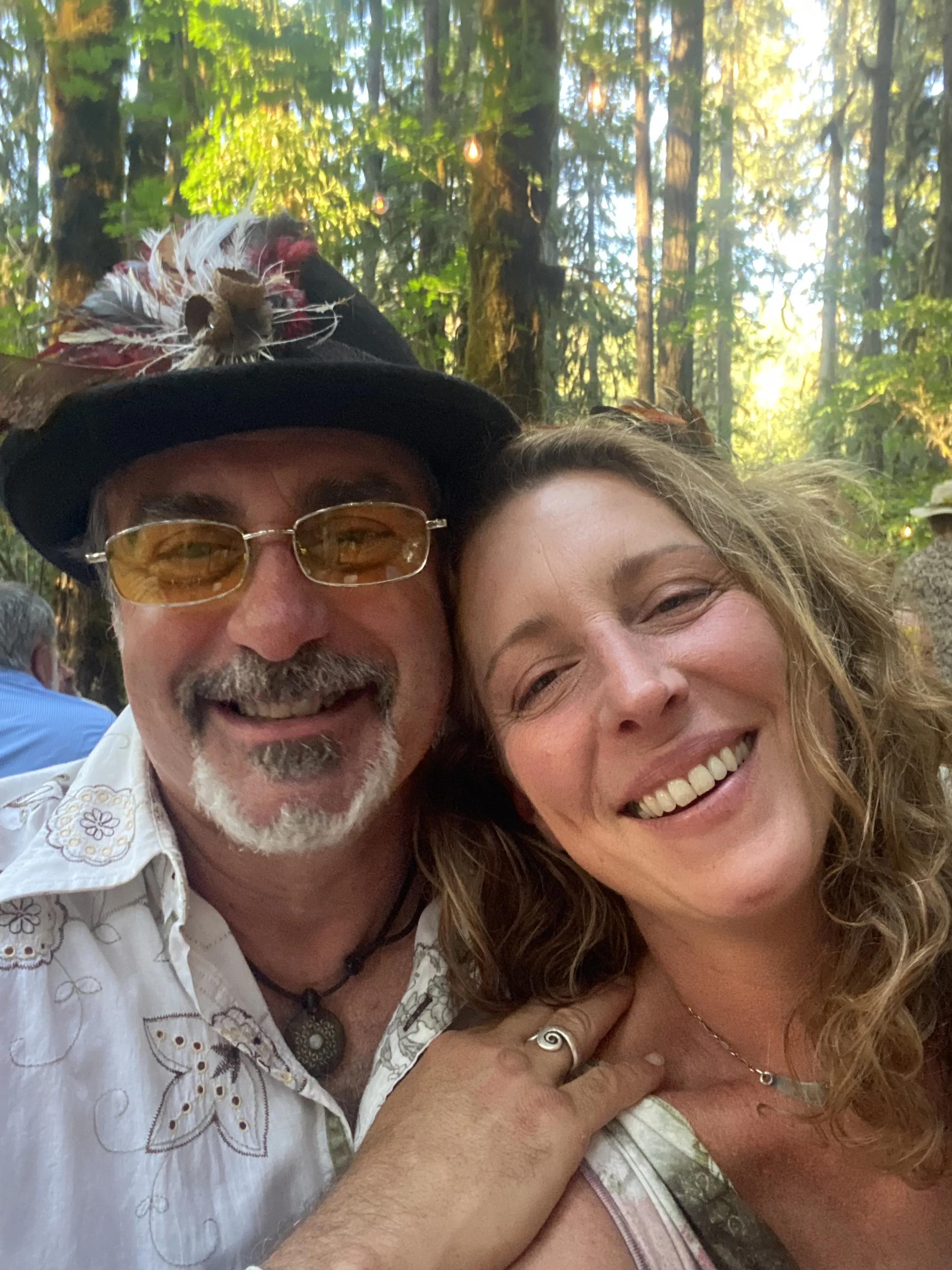 Two smiling people, a man wearing a black hat with feathers and glasses, and a woman with curly hair, in a forest setting during daytime.