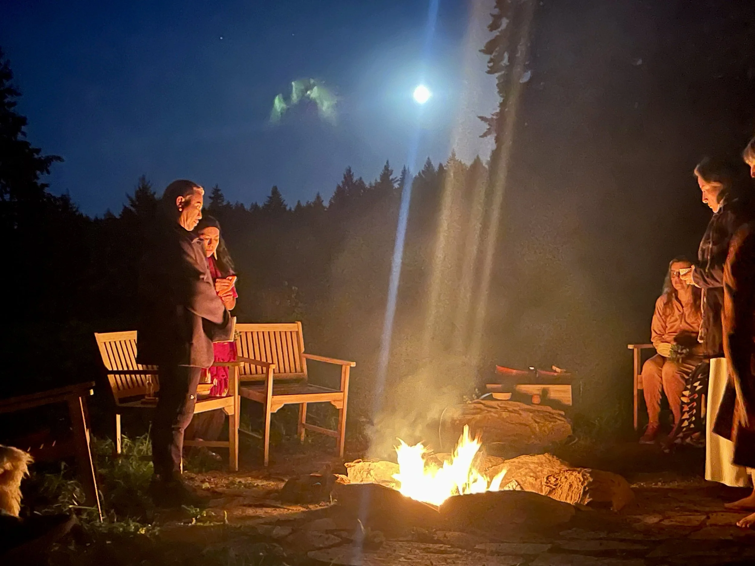 Group of people gathered around a campfire at night, with a full moon and starry sky overhead in a forested outdoor setting.