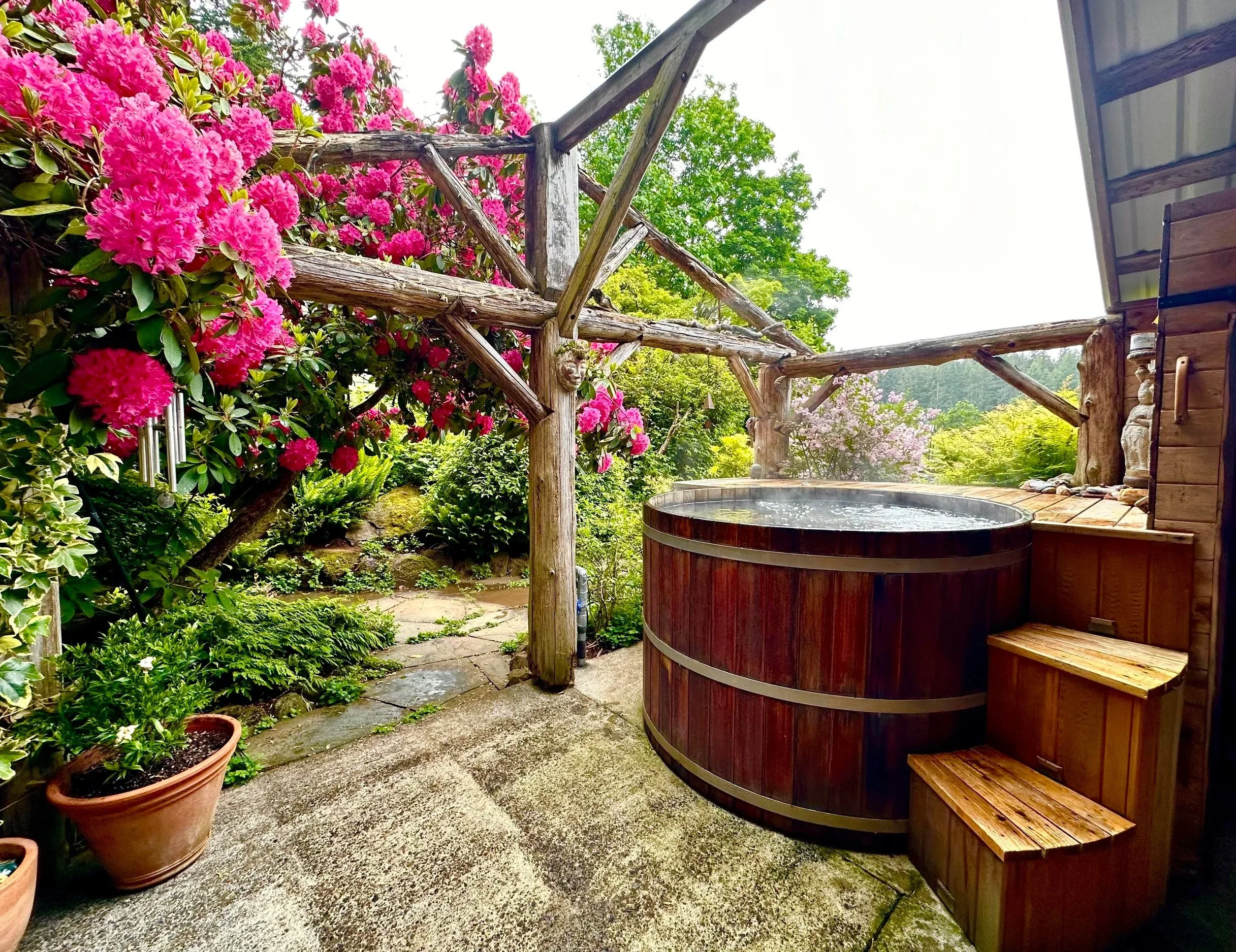 A cozy outdoor hot tub area surrounded by lush green and pink flowering bushes and plants, with wooden trim and outdoor stairs.