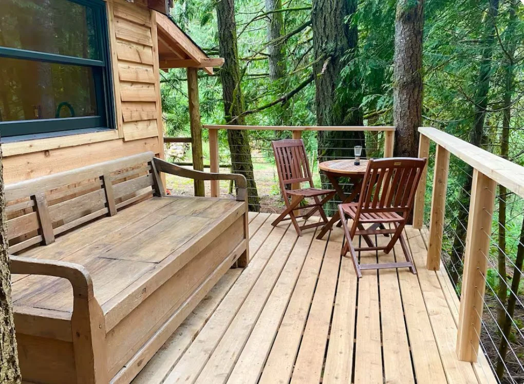 Wooden deck overlooking a forest, with a wooden bench, a round table, and two folding chairs, one with a glass of water on top.