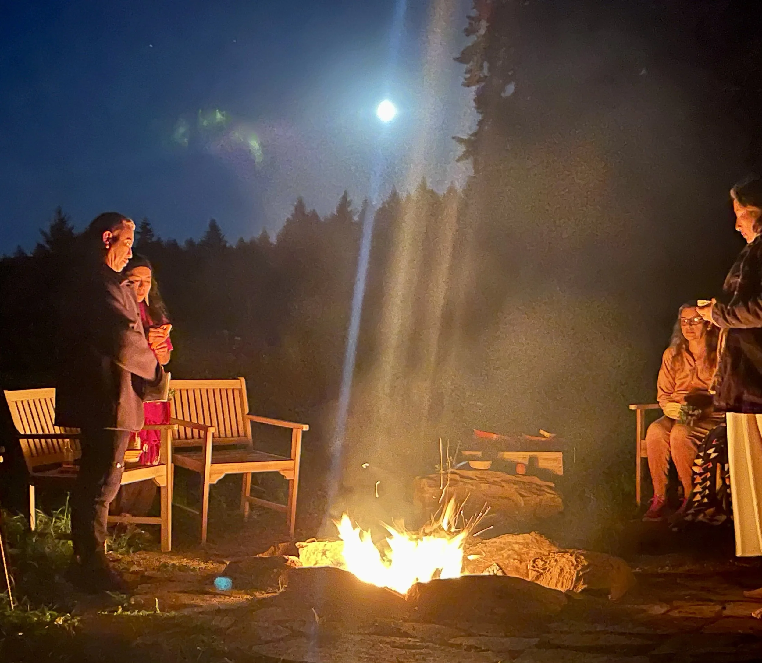Group of people gathered around a campfire at night, with the moon visible in the sky and trees in the background.
