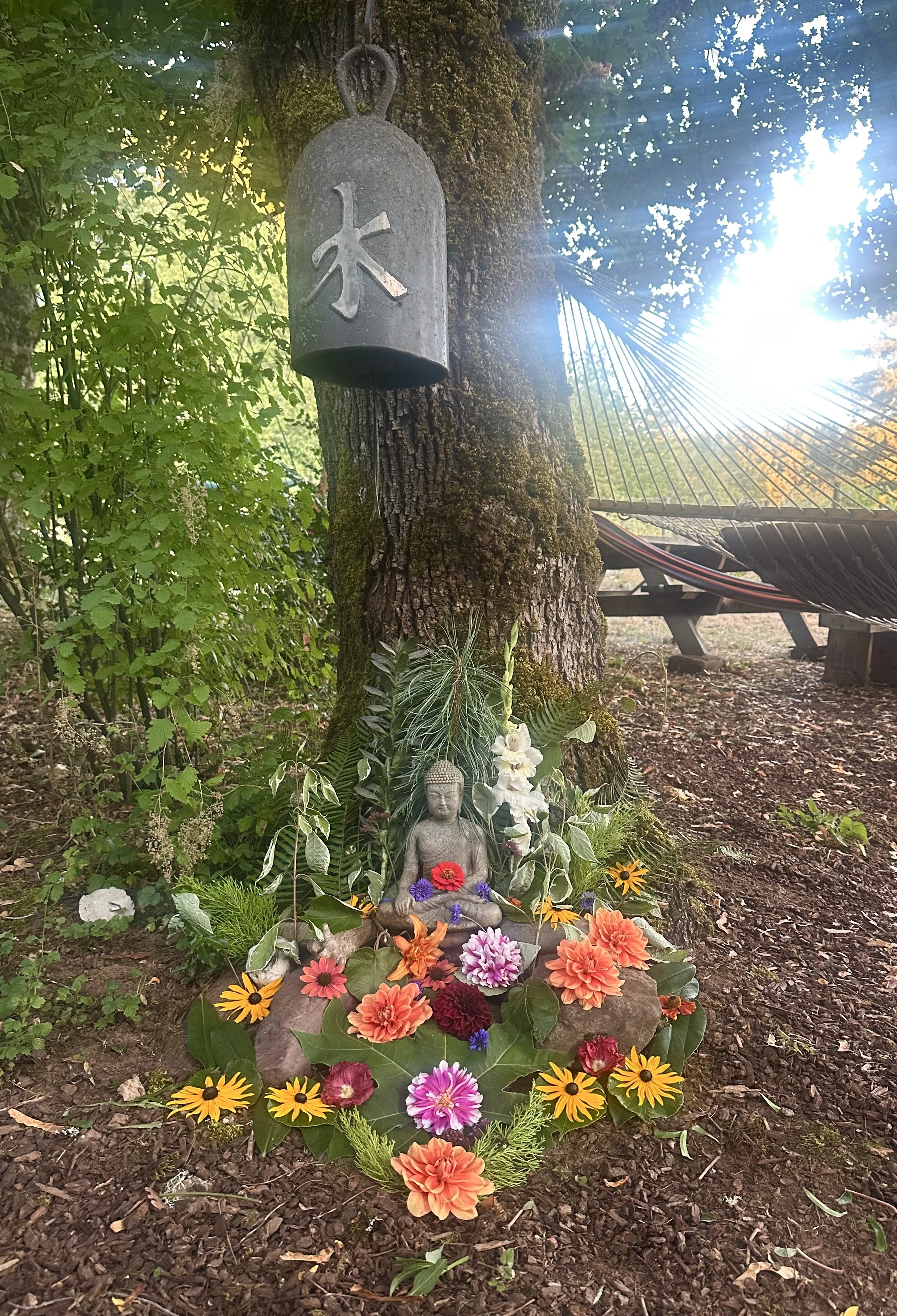 A small outdoor shrine scene with a stone Buddha statue surrounded by colorful flowers and various green plants, situated at the base of a moss-covered tree. A metal bell with a Japanese character hangs from the tree above, and in the background, gre