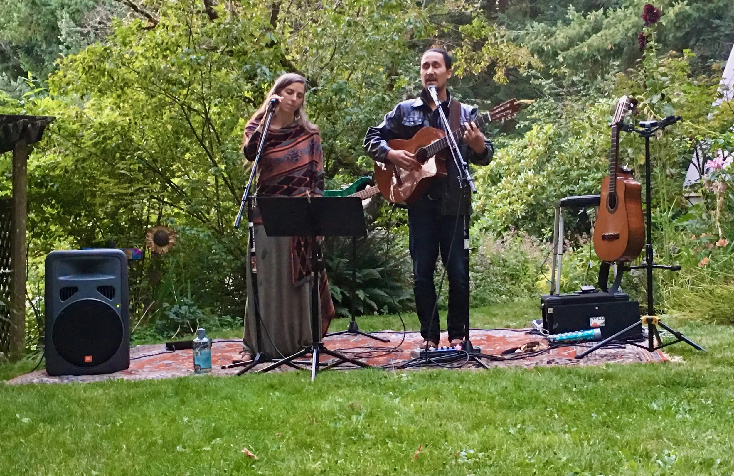 Two musicians performing outdoors on a grassy area surrounded by trees and bushes, with one playing guitar and singing, the other holding a microphone, with additional guitars and equipment on stands nearby.