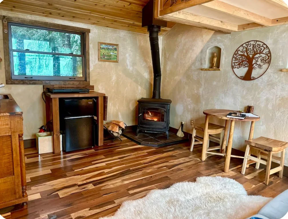 Cozy living room with wood flooring, a wood stove, a small refrigerator, a window with view of trees, a fluffy white rug, wooden furniture including a table and stools, wall decorations including a metal tree art, a small statue in a niche, and a pai
