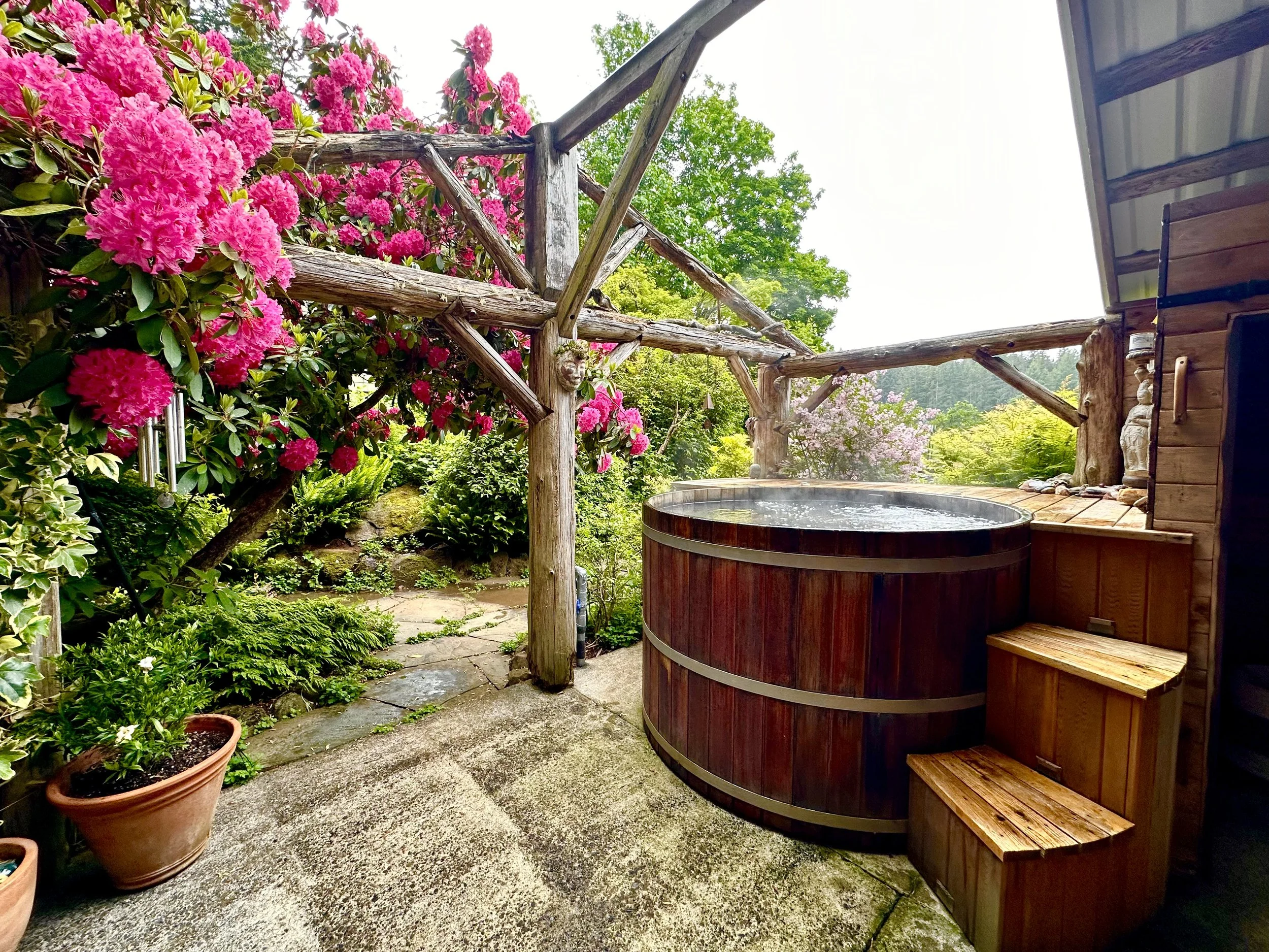 Outdoor garden with pink flowering bushes, potted plants, a wooden hot tub, and a rustic wooden railing under a metal roof.