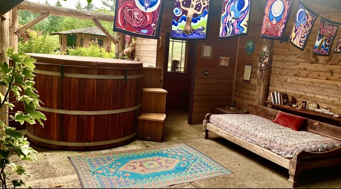 A cozy outdoor space with a hot tub, colorful hanging artwork, a vintage-style sofa with a red pillow, a patterned rug, and various decorative items on a shelf against wooden walls.