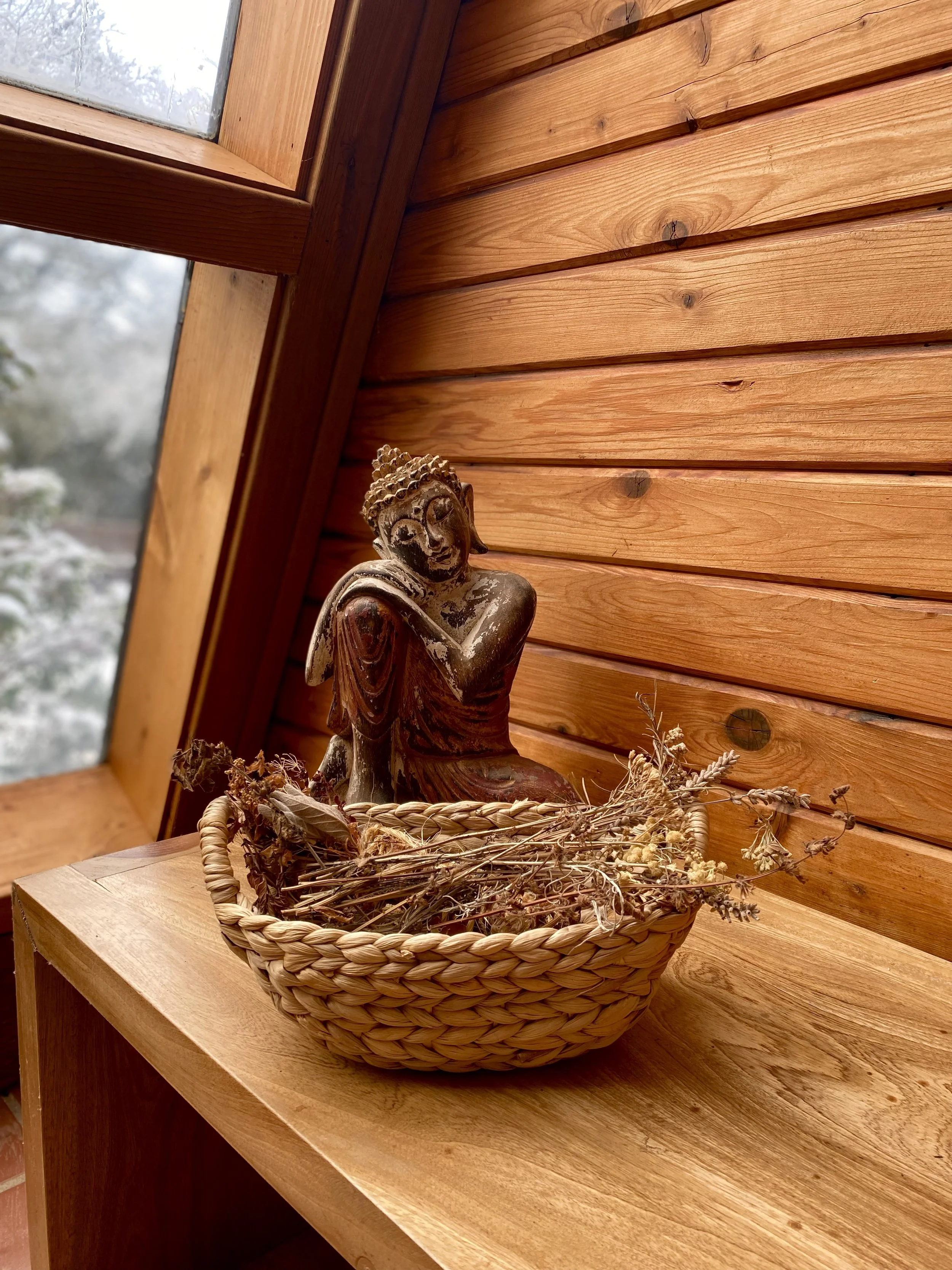 A wooden Buddha statue sitting in a woven basket filled with dried herbs and flowers, placed on a wooden surface inside a room with wood-paneled walls and a sloped ceiling with a window.