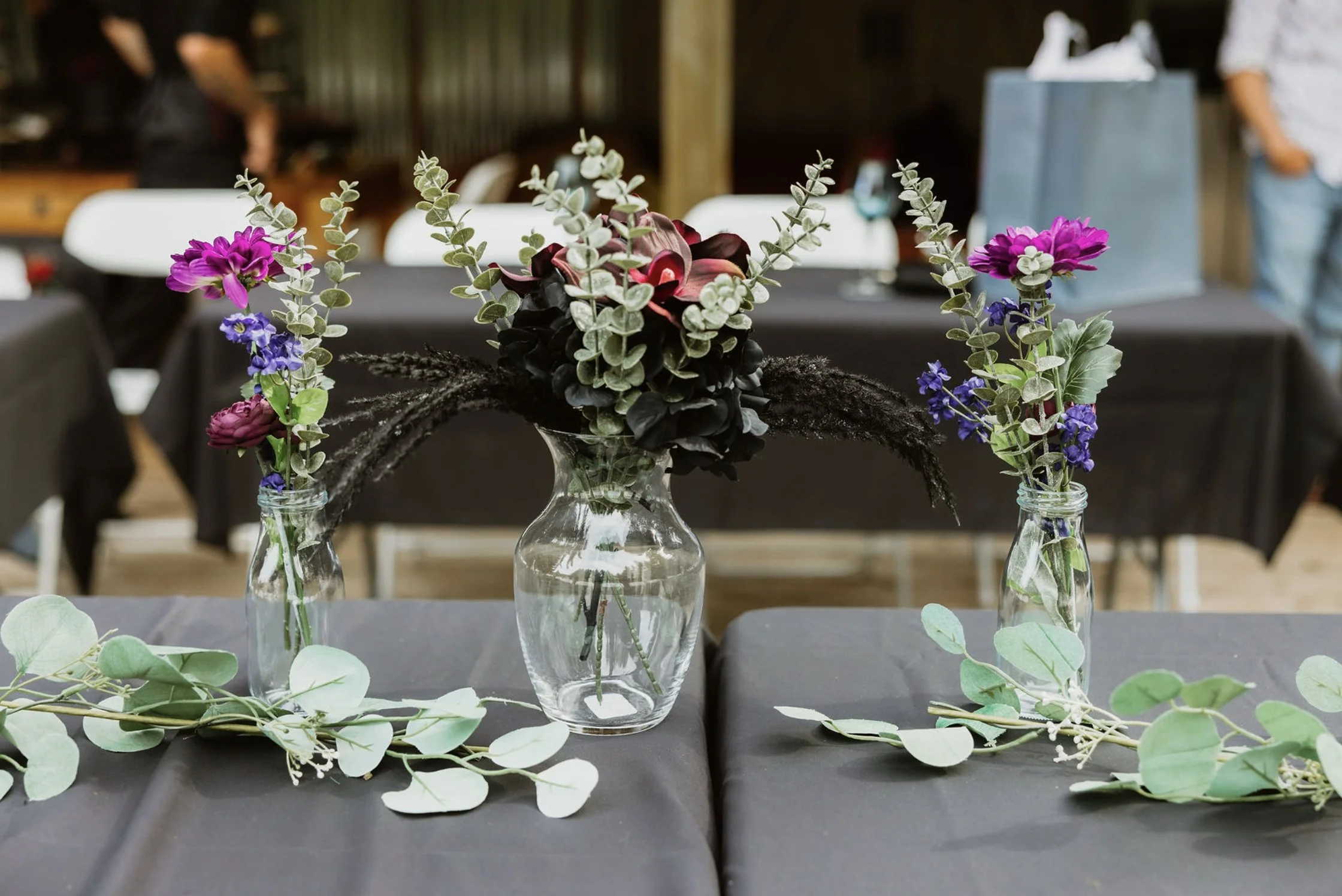 Floral arrangement with purple, pink, and black flowers in a vase, flanked by two smaller vases with purple and blue flowers, placed on a black tablecloth in an indoor setting.