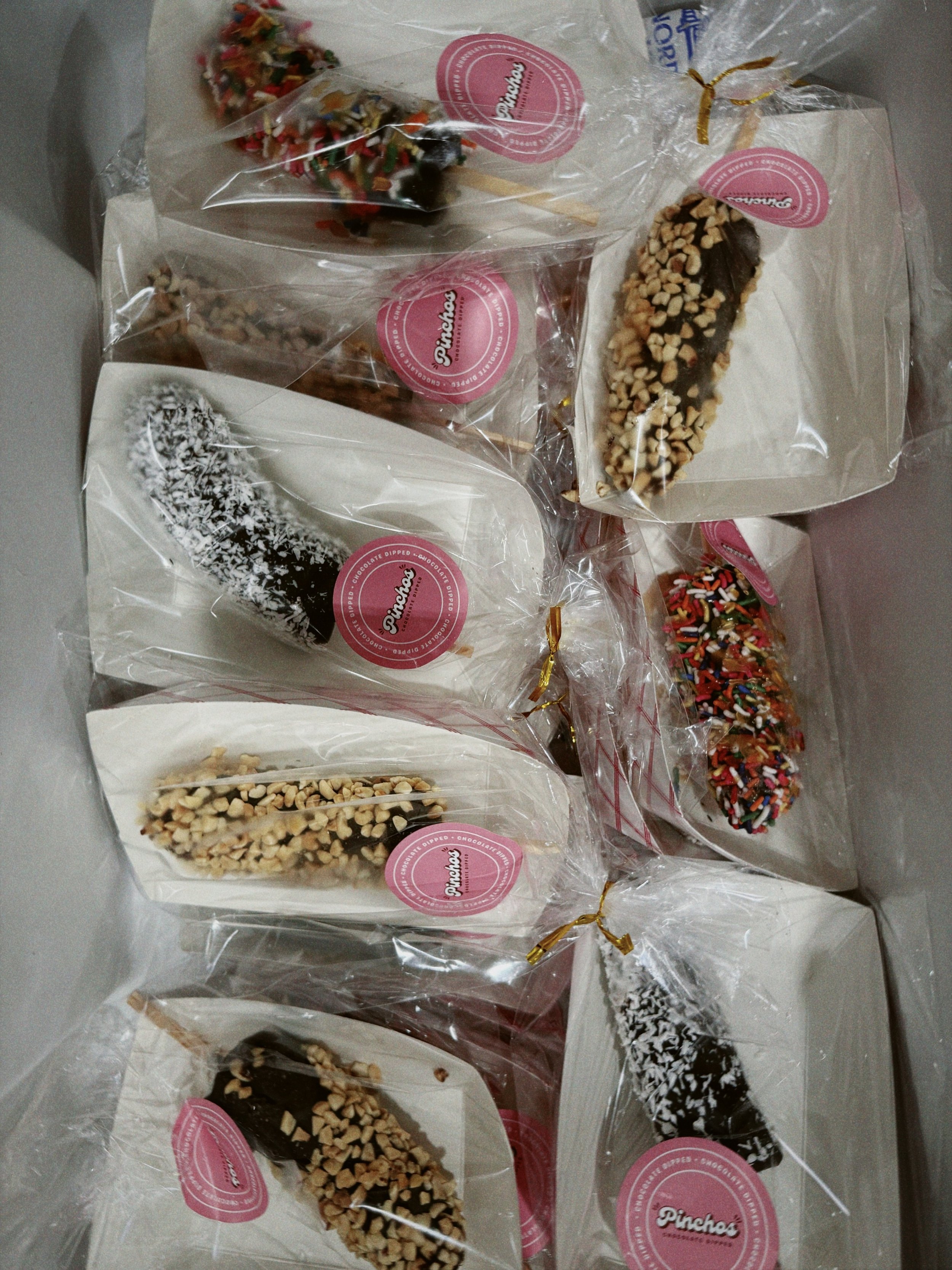 A collection of assorted chocolate-covered treats wrapped in clear plastic with pink Pinch's labels, including some topped with sprinkles, chopped nuts, shredded coconut, and colorful sprinkles.