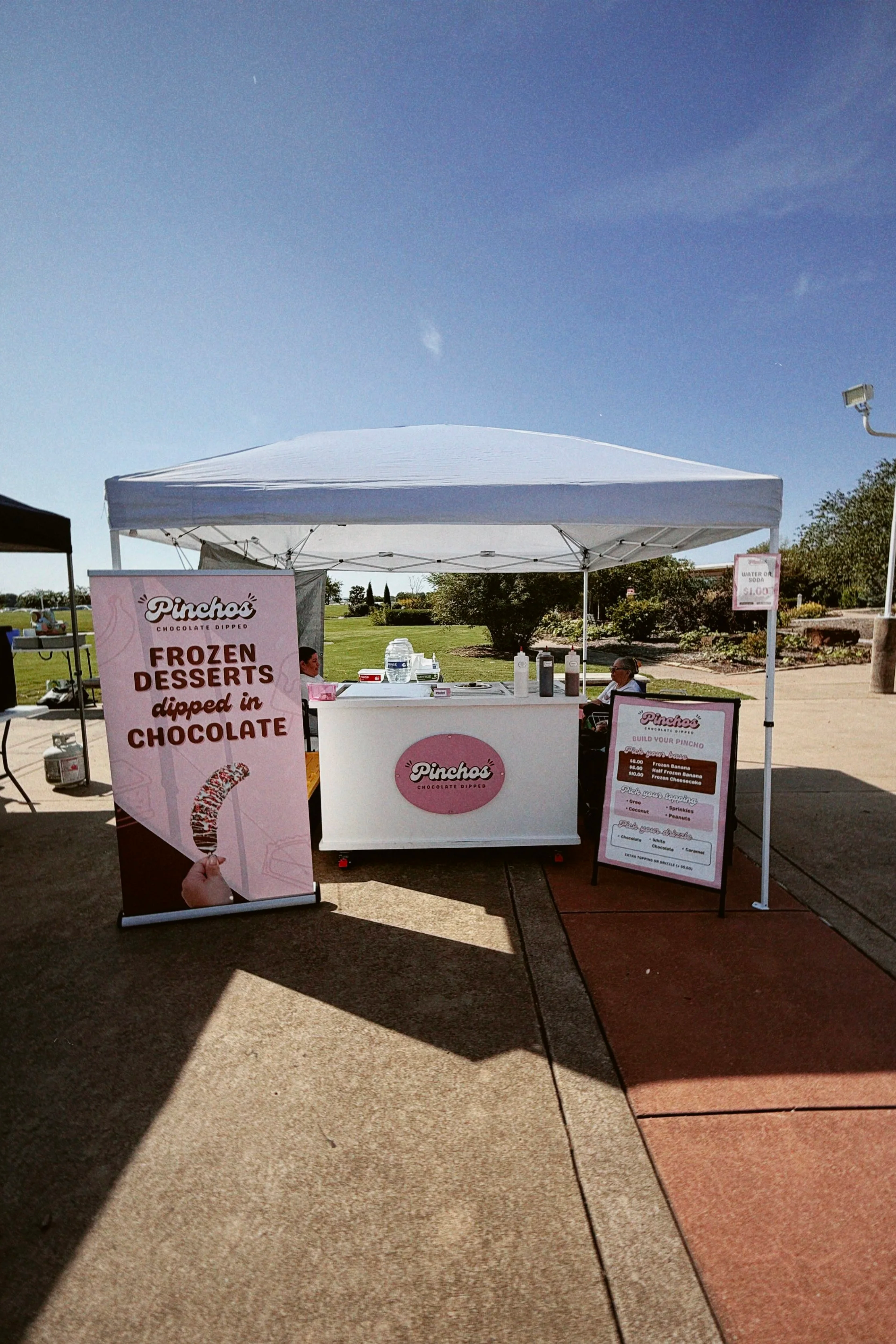 A small outdoor dessert stand named "Pincho's" selling frozen desserts dipped in chocolate, with a pink sign, located under a white canopy tent. There are signs and a menu board displayed in front, with a grassy park in the background.