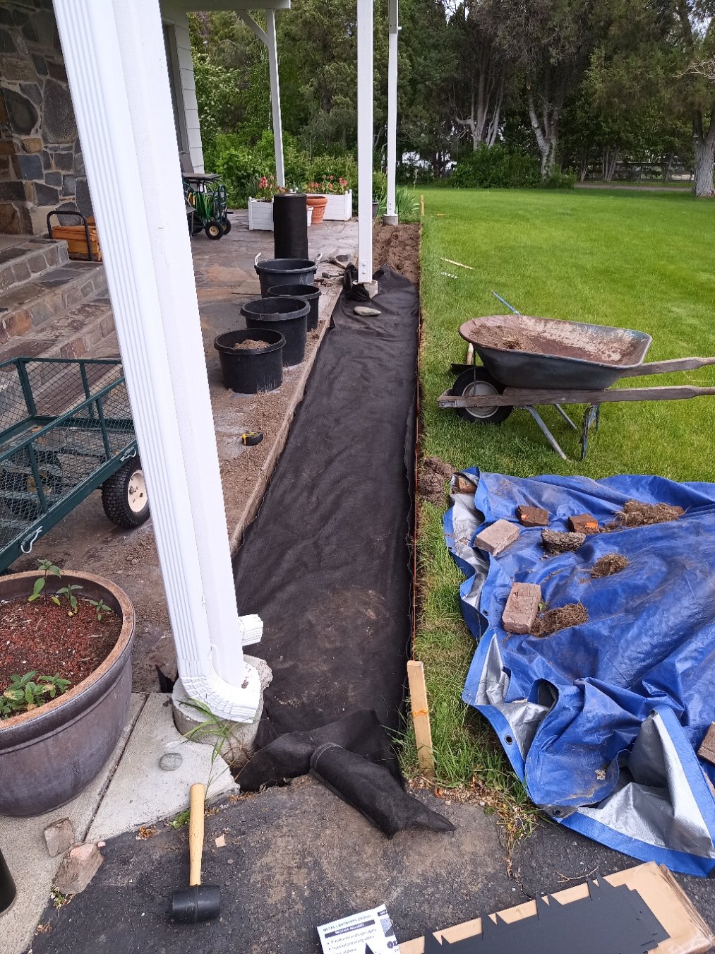 Landscape project with black fabric lining a trench, surrounded by potted plants, gardening tools, and a wheelbarrow, with a patio and grass lawn in the background.